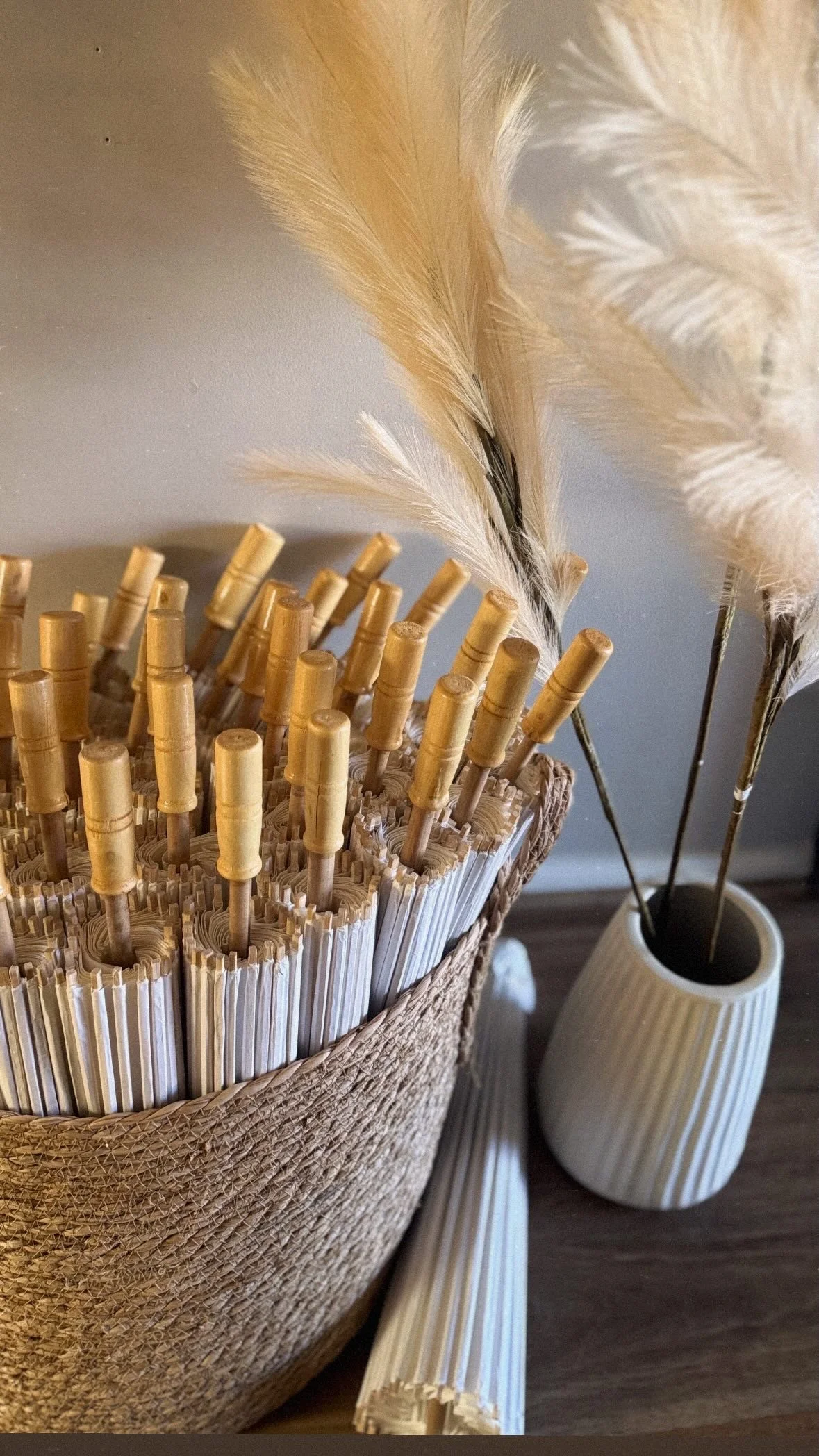 A woven basket filled with rolled paper straws with bamboo-like handles, placed on a wooden surface. In the background, there is a white ribbed vase holding tall, fluffy pampas grass.