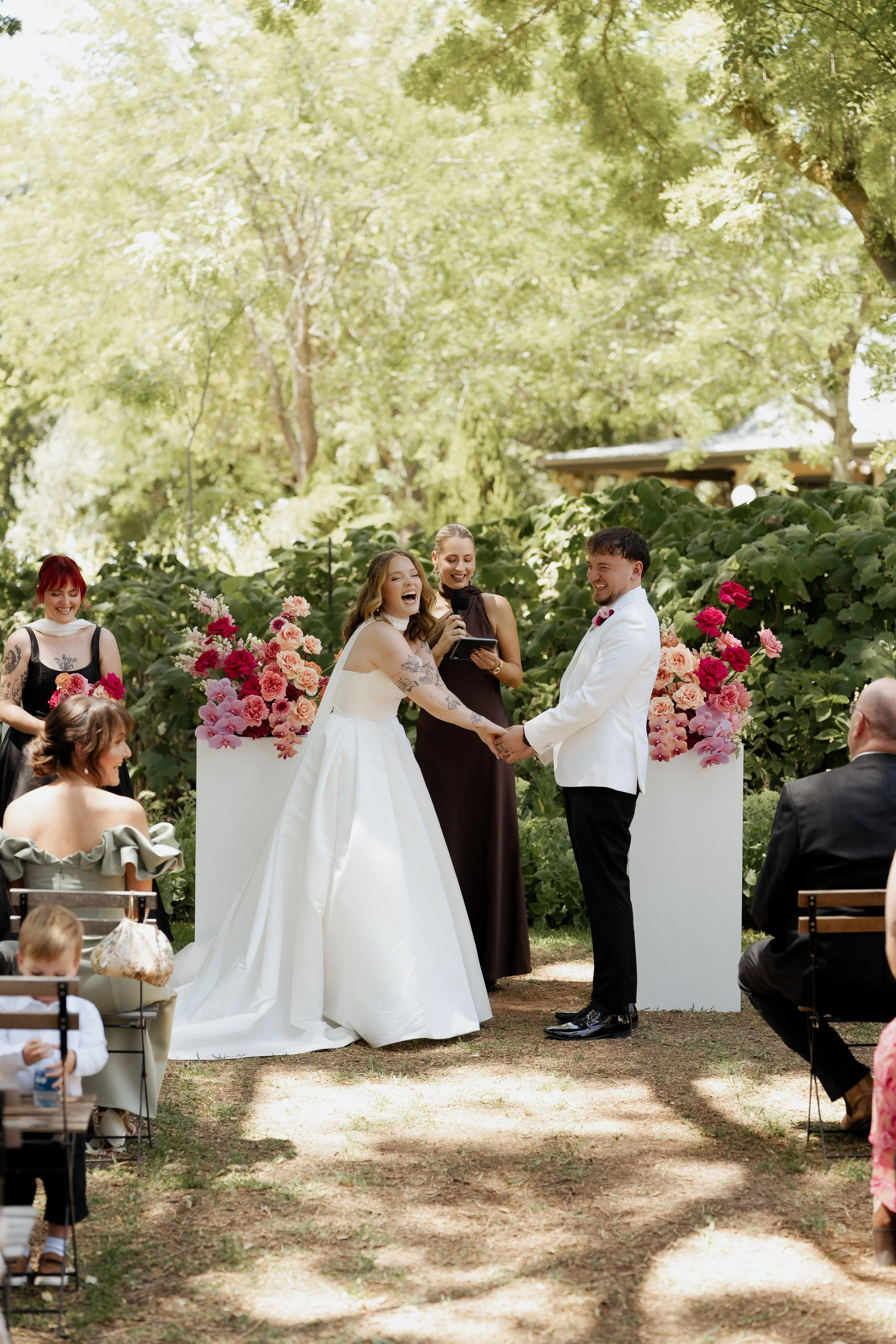 A wedding ceremony taking place outdoors with a bride and groom holding hands and smiling, surrounded by friends and family, in a lush green garden with trees and floral arrangements.