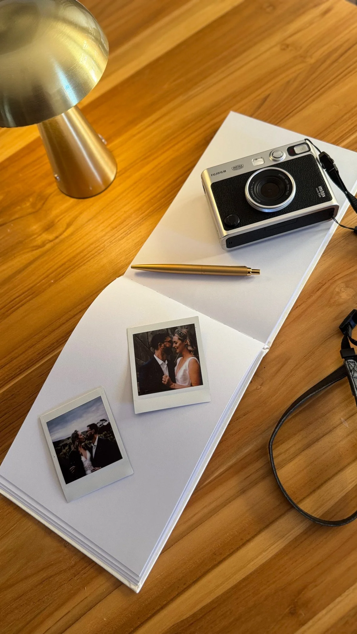 A wooden table with a white notepad, two Polaroid photos of a couple, a gold pen, a small silver digital camera, and a black strap.