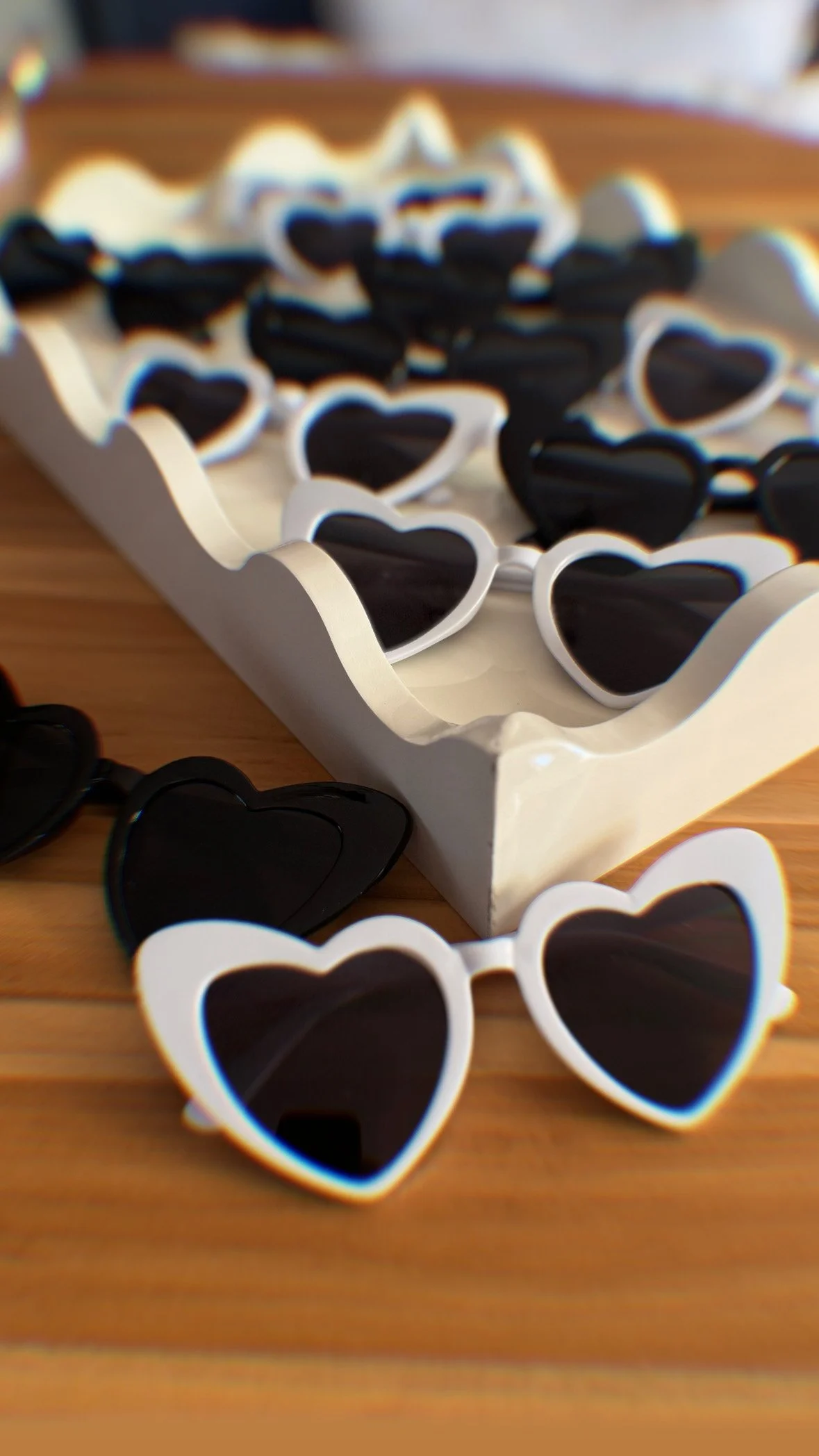 A white tray holding numerous black and white heart-shaped sunglasses on a wooden table.