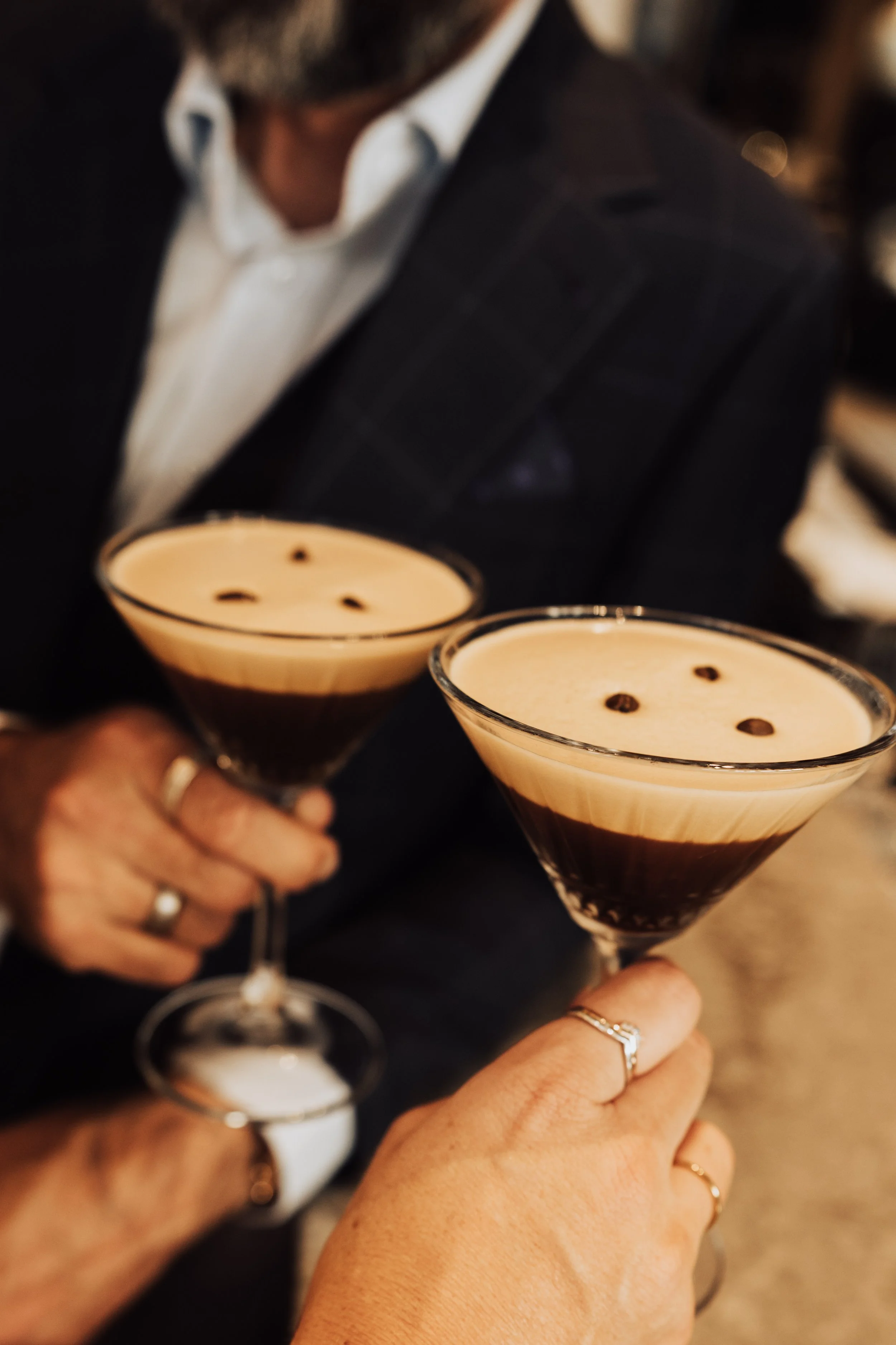 Two people holding cocktail glasses with layered drinks topped with foam and chocolate dots, dressed in formal attire.