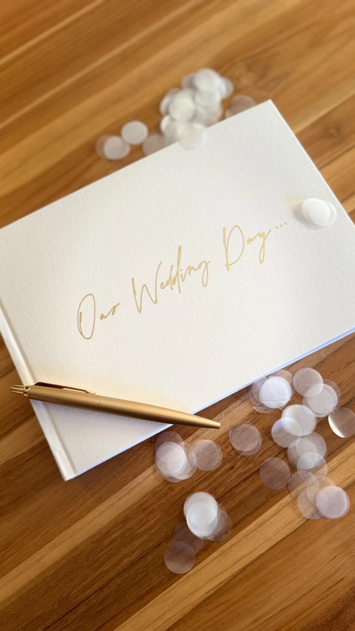 A wedding invitation card with gold pen on wooden table, surrounded by white and translucent confetti.