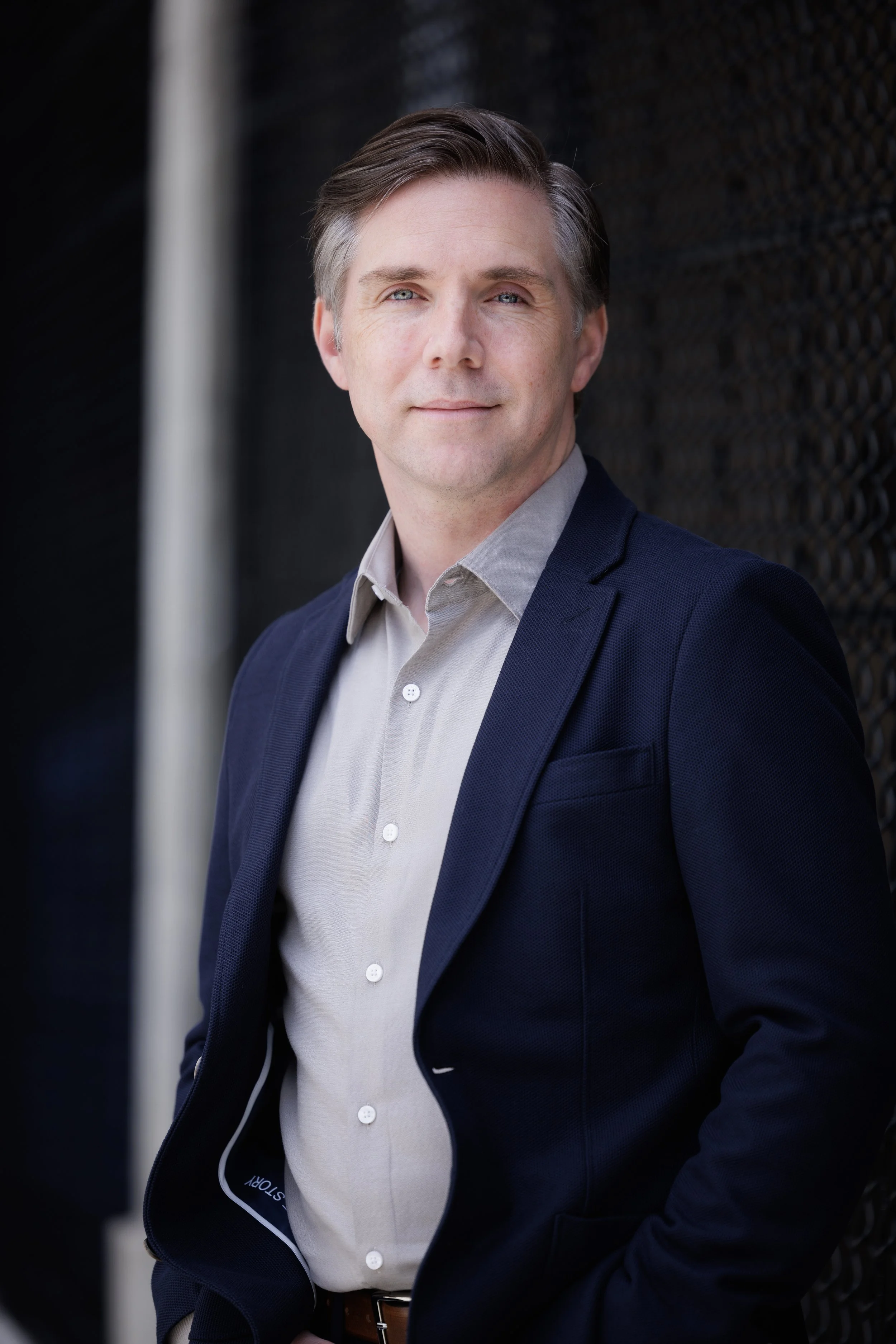 A man with light brown hair and blue eyes, wearing a gray collared shirt and a dark blue blazer, standing outdoors against a black textured wall.