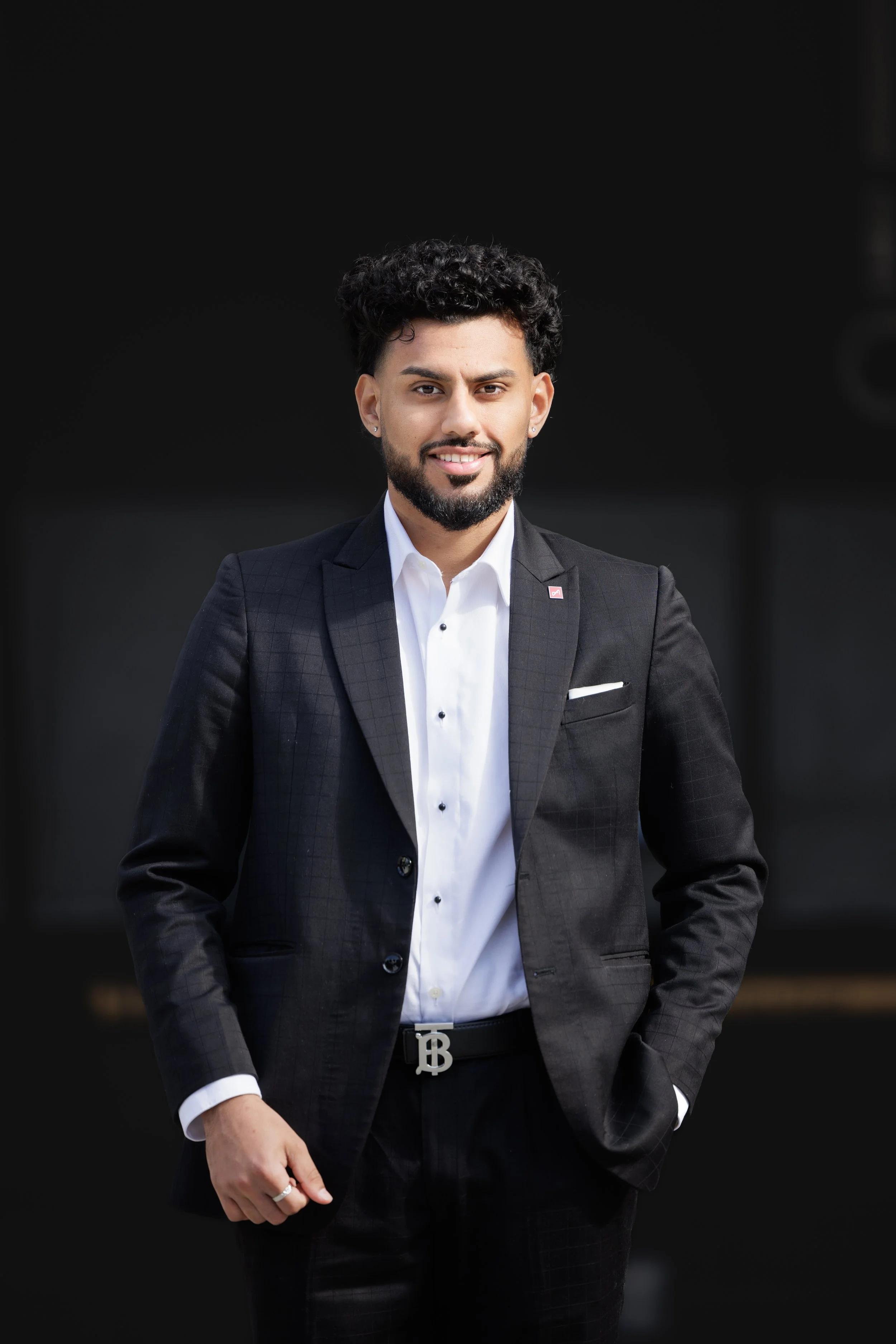 Portrait of a man in a black suit with a white shirt, smiling against a dark background.