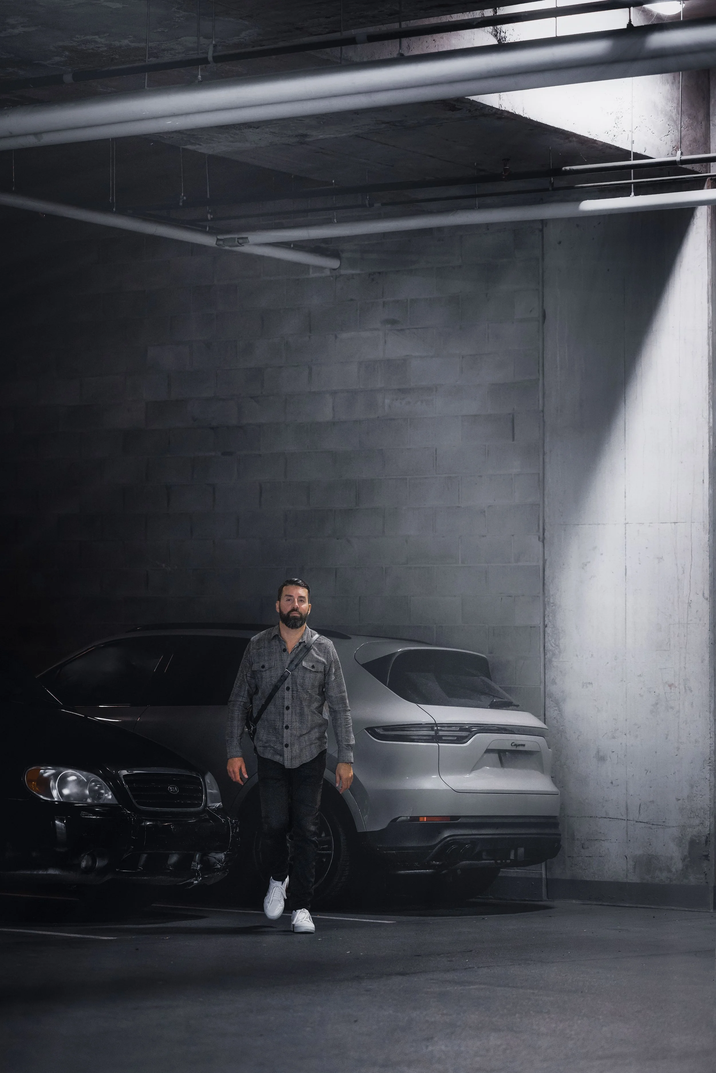 A man with a beard and gray jacket walking in an underground parking garage next to two parked cars, one black and one gray.