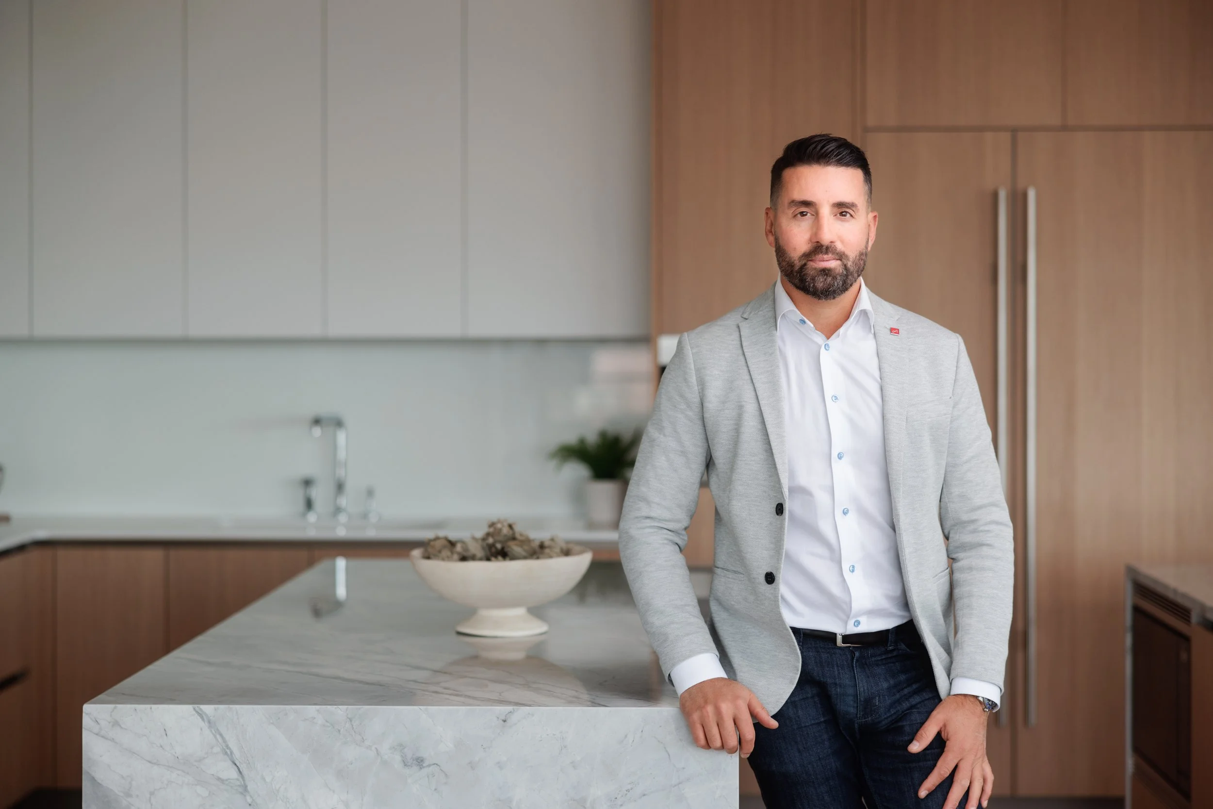 A man with a beard and short dark hair in business casual attire standing in a modern kitchen with a marble island and wooden cabinets.
