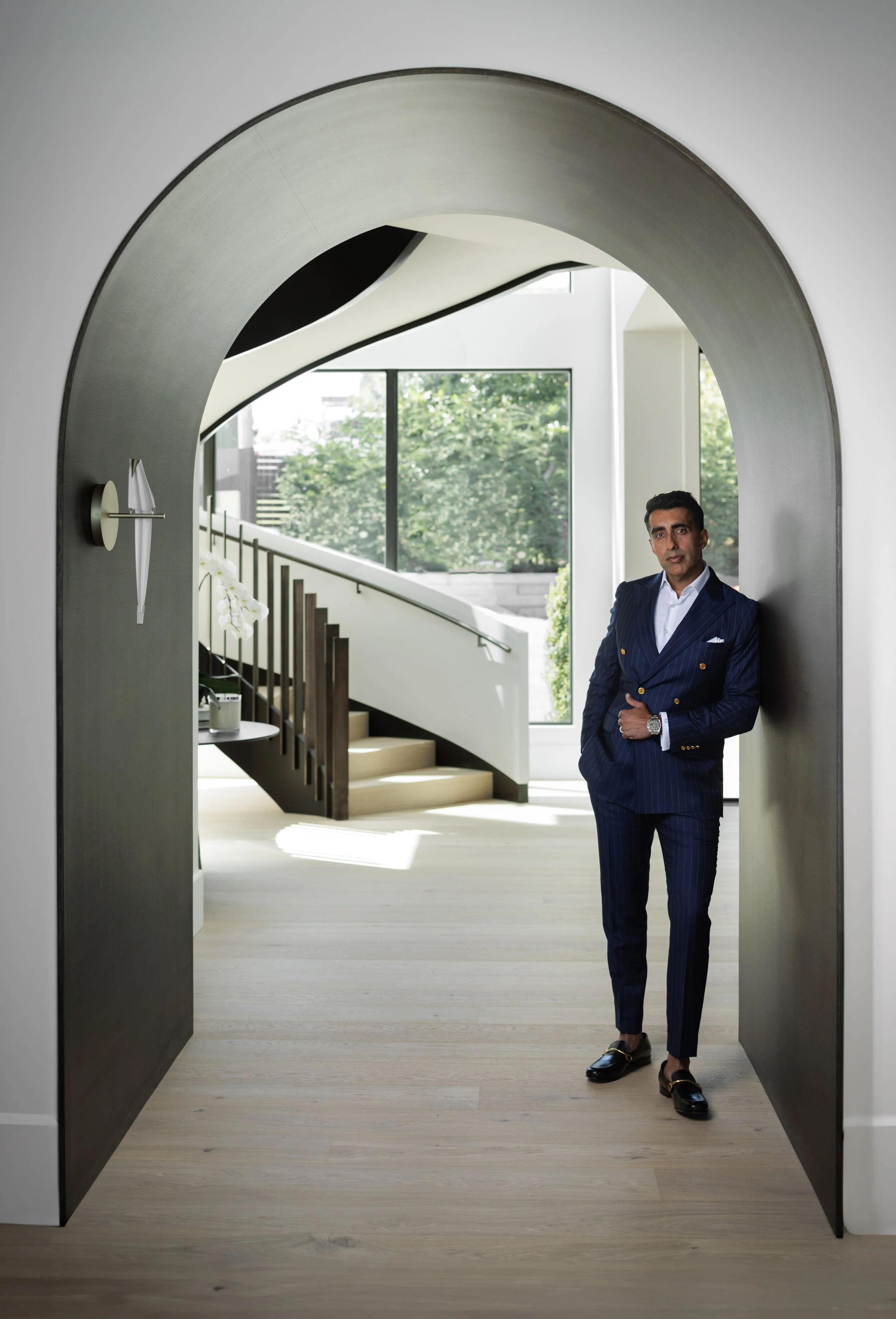 A man in a navy blue pinstripe suit stands in a doorway with large black doors, inside a modern, well-lit home with large windows, stairs, and greenery outside.