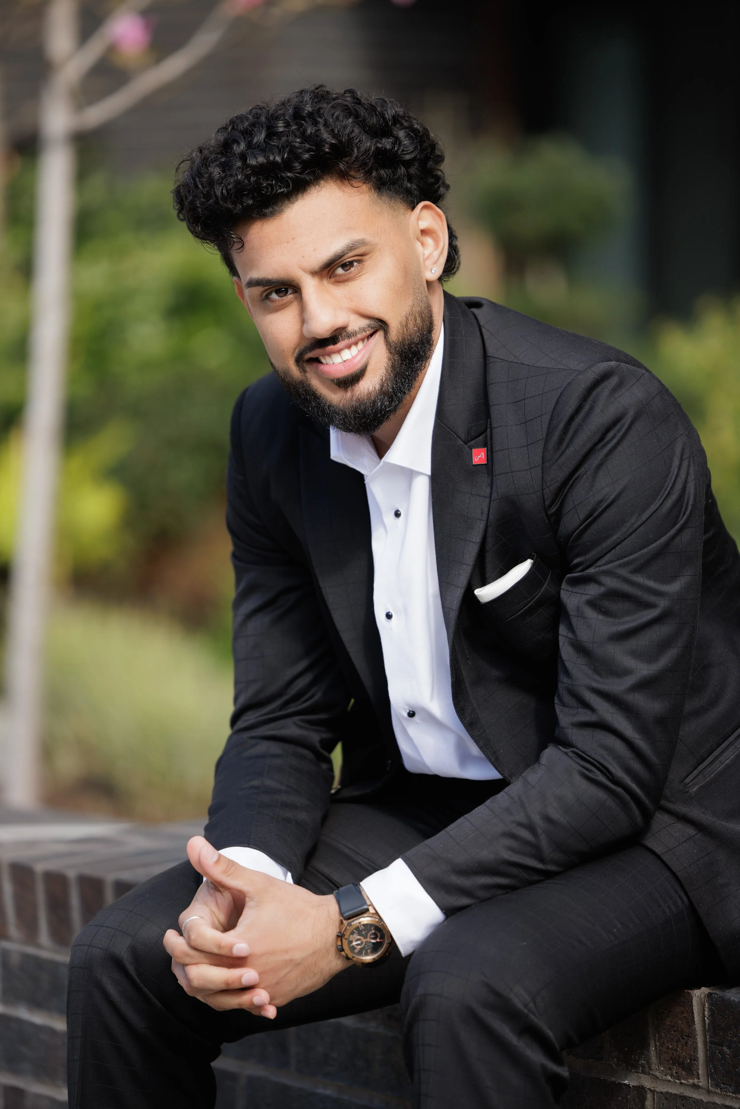 A smiling man in a black suit with a white dress shirt sitting outdoors on a brick ledge, with a blurred green background.