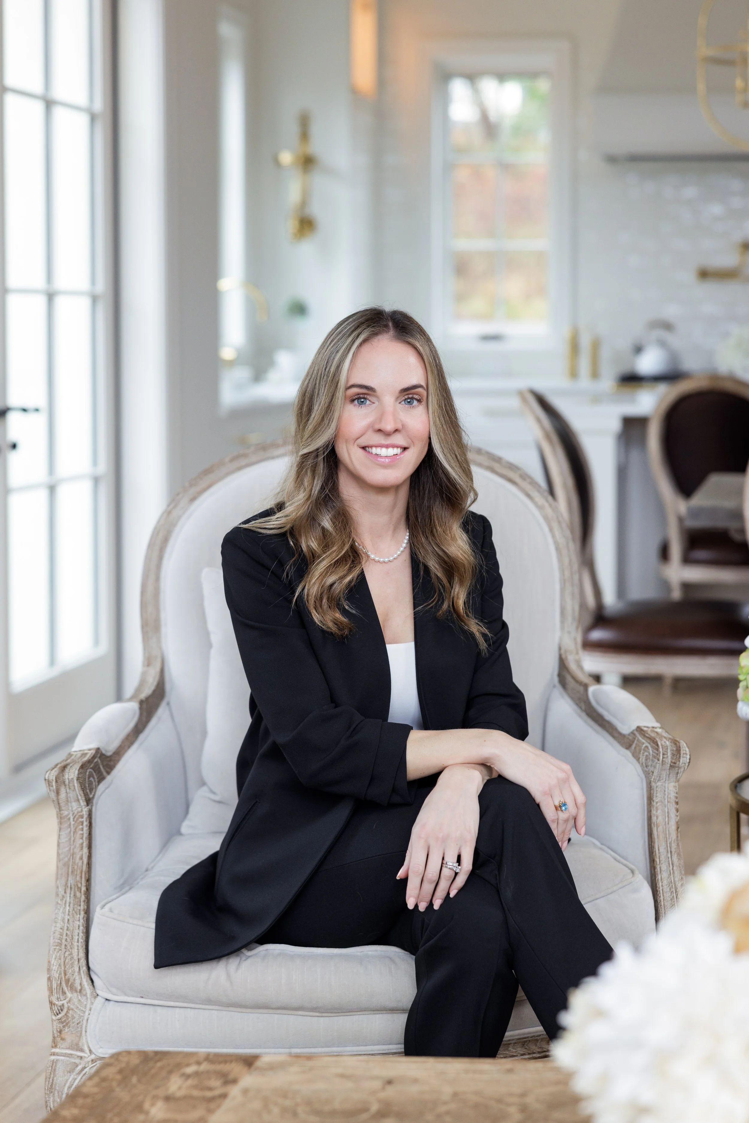 A woman with long wavy hair, wearing a black blazer, white top, and pearl necklace, sits on a white upholstered armchair in a bright, elegant kitchen or dining area, smiling at the camera.