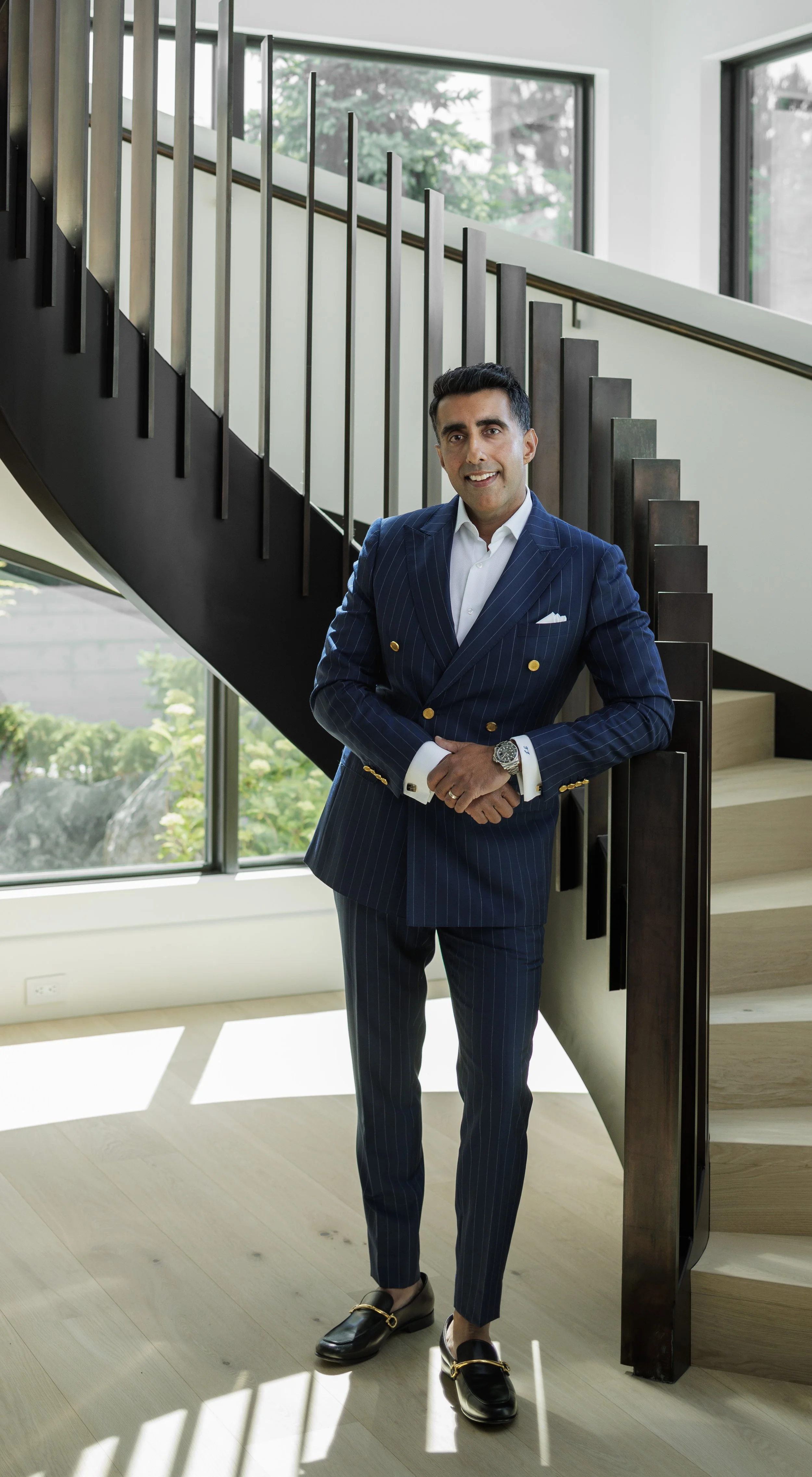 A man in a navy blue pinstripe suit standing inside a modern house, smiling, with large windows and a staircase in the background.