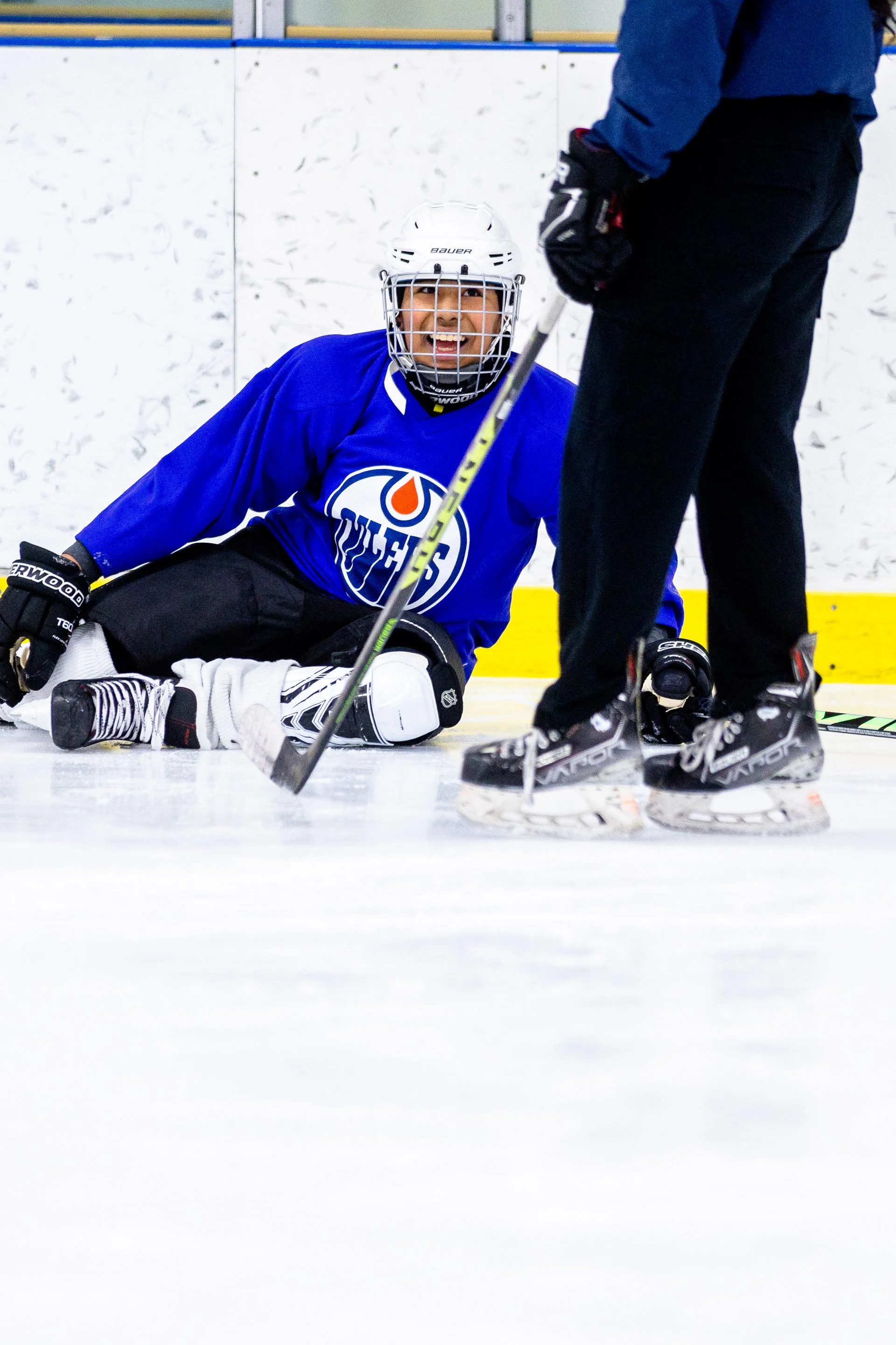 A young hockey player sitting on the ice smiles up at a coach out of frame, wearing a blue Oilers practice jersey.