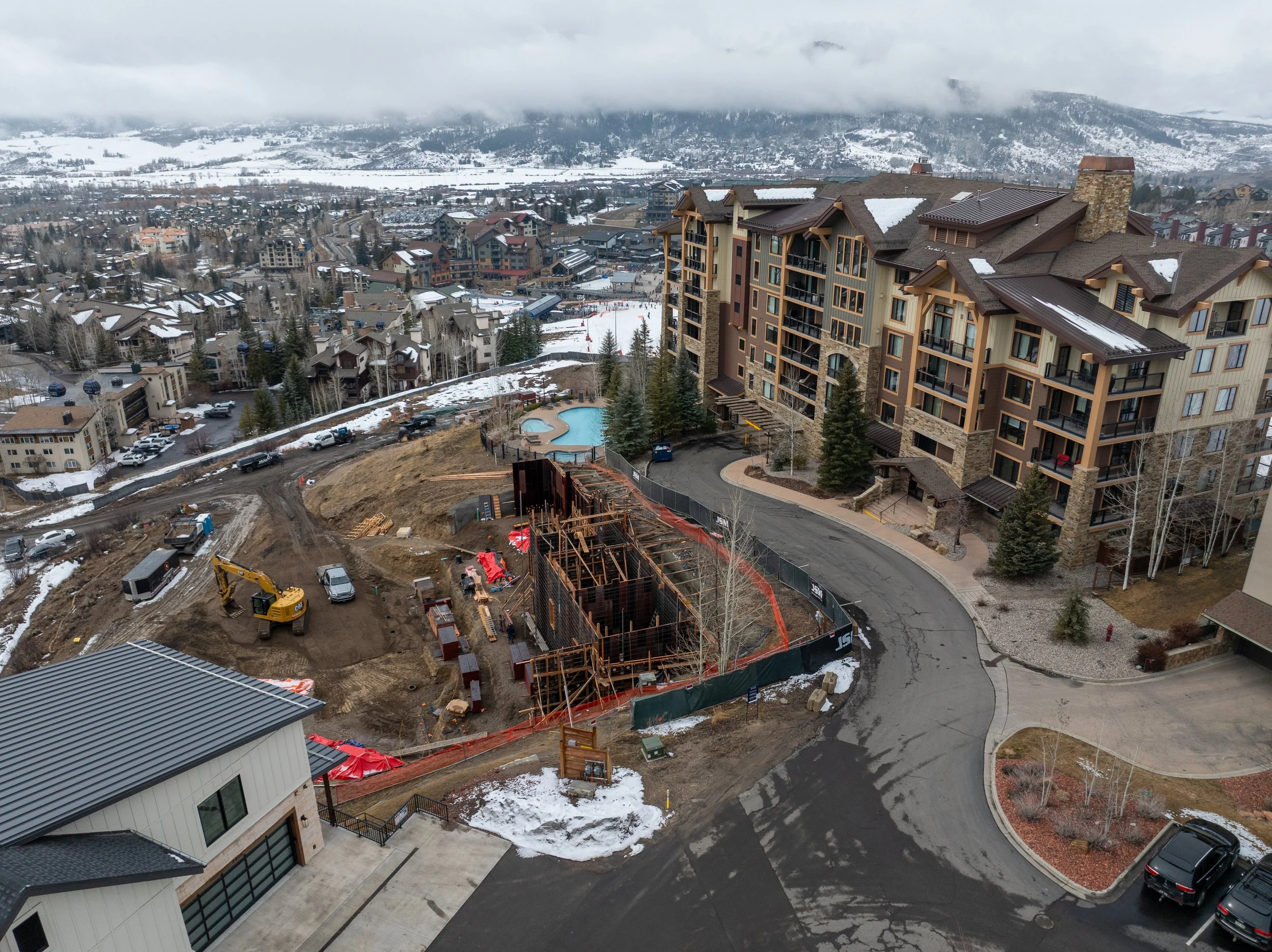 Nighttime rendering of modern mountain lodge with stone and wood exterior, glass balconies, and multiple gabled roofs, set on a snowy hillside with a mountain backdrop.