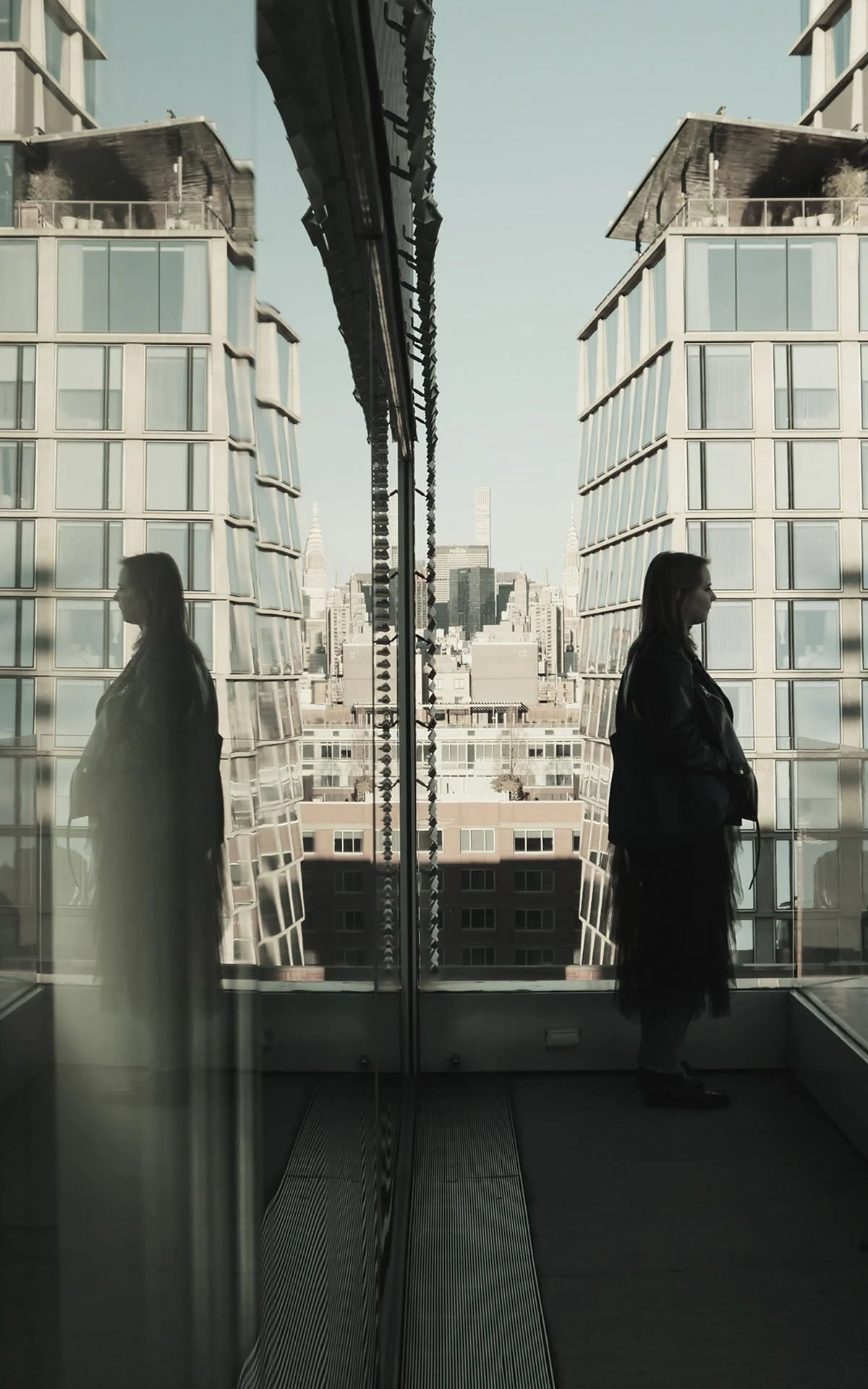 A woman stands on a balcony inside a building, reflected in a glass wall, with city buildings including the Empire State Building in the background.