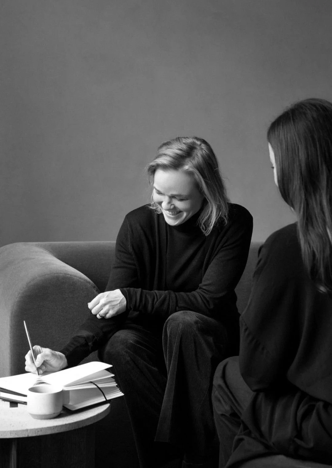 Two women sitting on a sofa, smiling and laughing during a conversation. One woman is taking notes on a notepad, and there are notebooks and a mug on a small table in front of them.