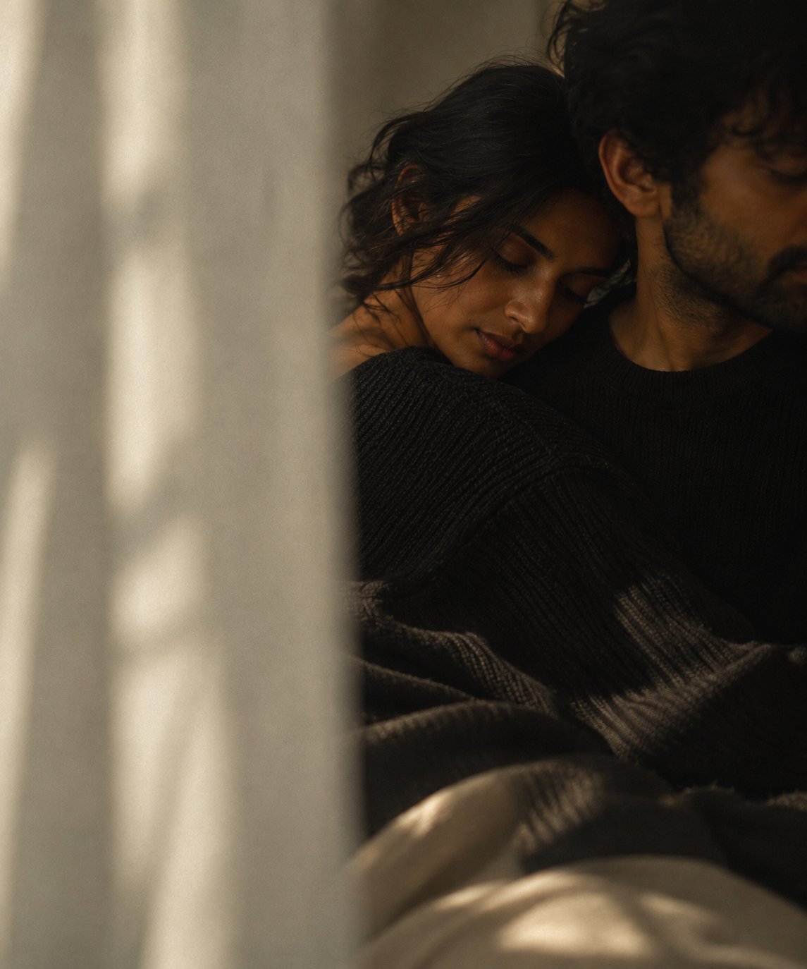 A woman resting her head on a man's shoulder with her eyes closed, sitting close together in a cozy, dimly lit setting.