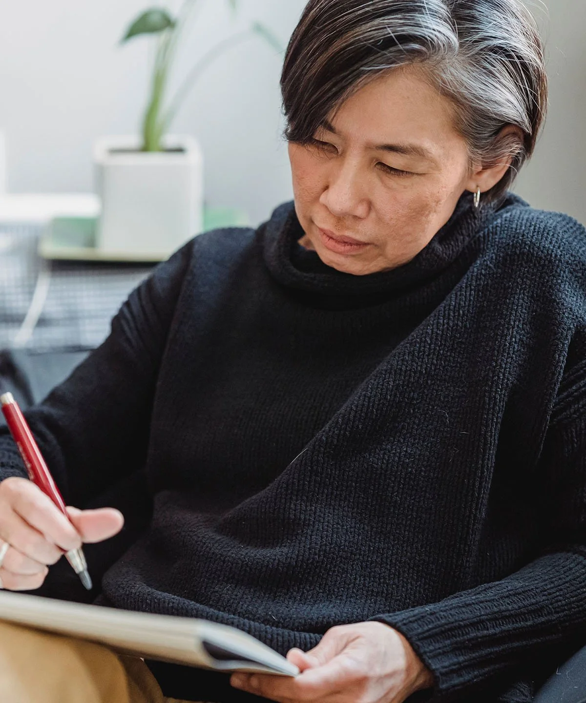 A woman with short dark hair and earrings, wearing a black sweater, is sitting at a desk, writing in a notebook with a red pen.