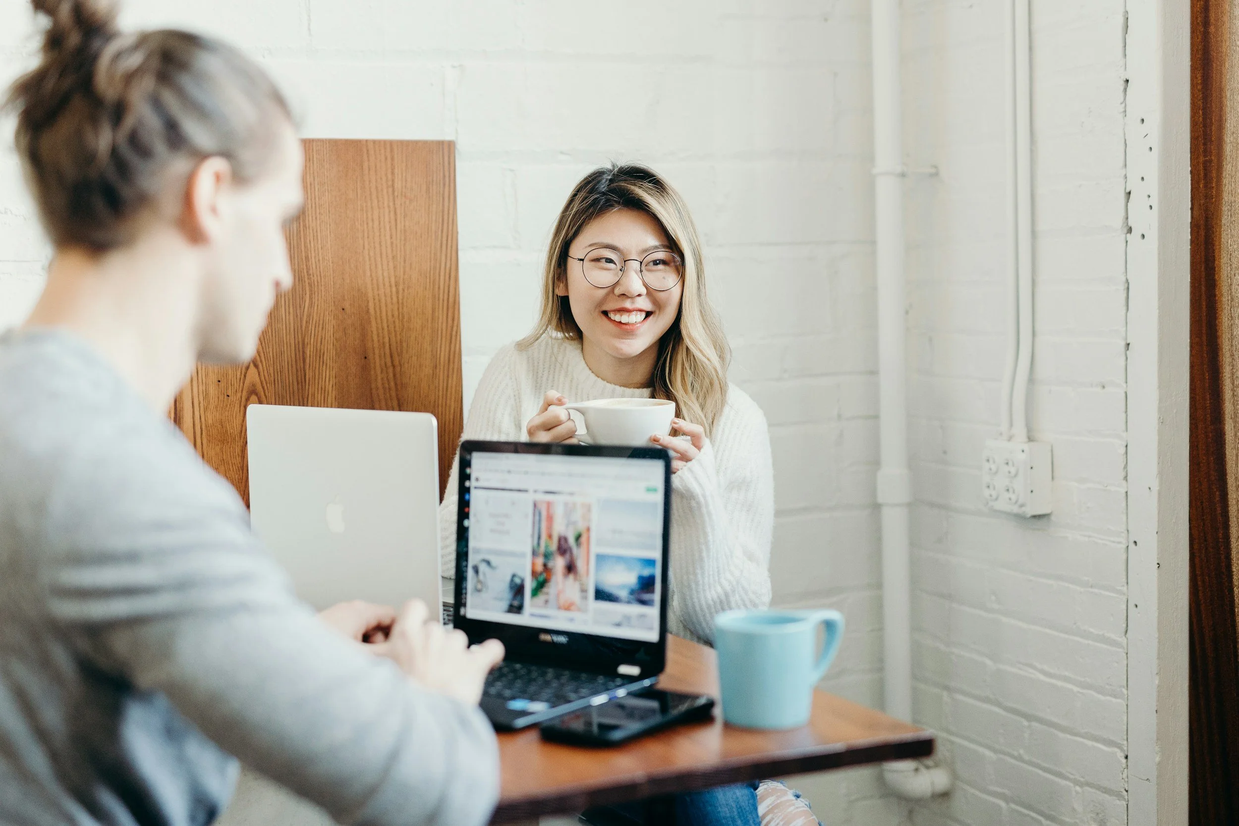 A woman with glasses smiling and holding a white mug while sitting at a table with a man using a laptop in a cozy, white-painted room.