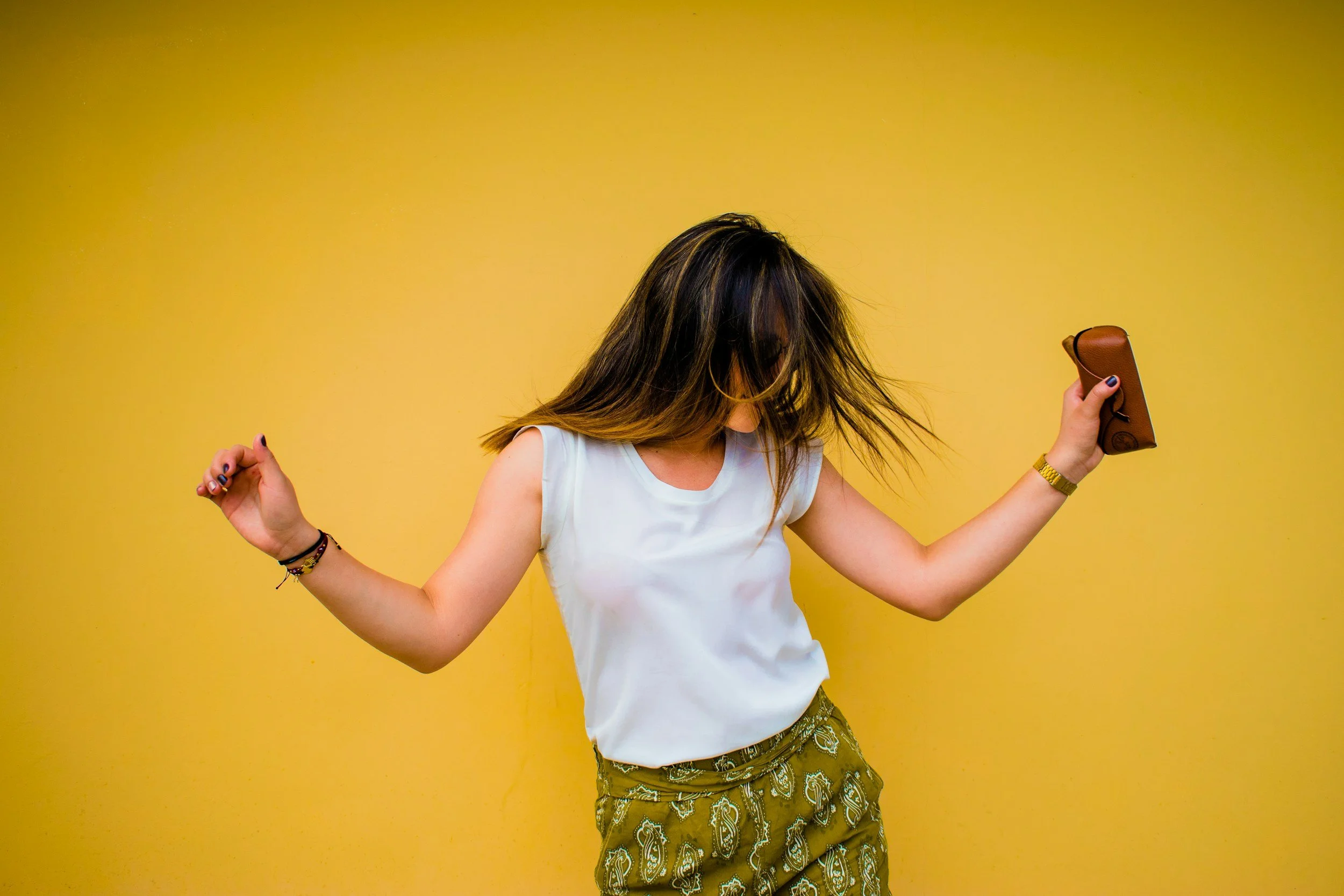 Woman dancing with hair flying, holding a brown wallet, against a yellow wall.
