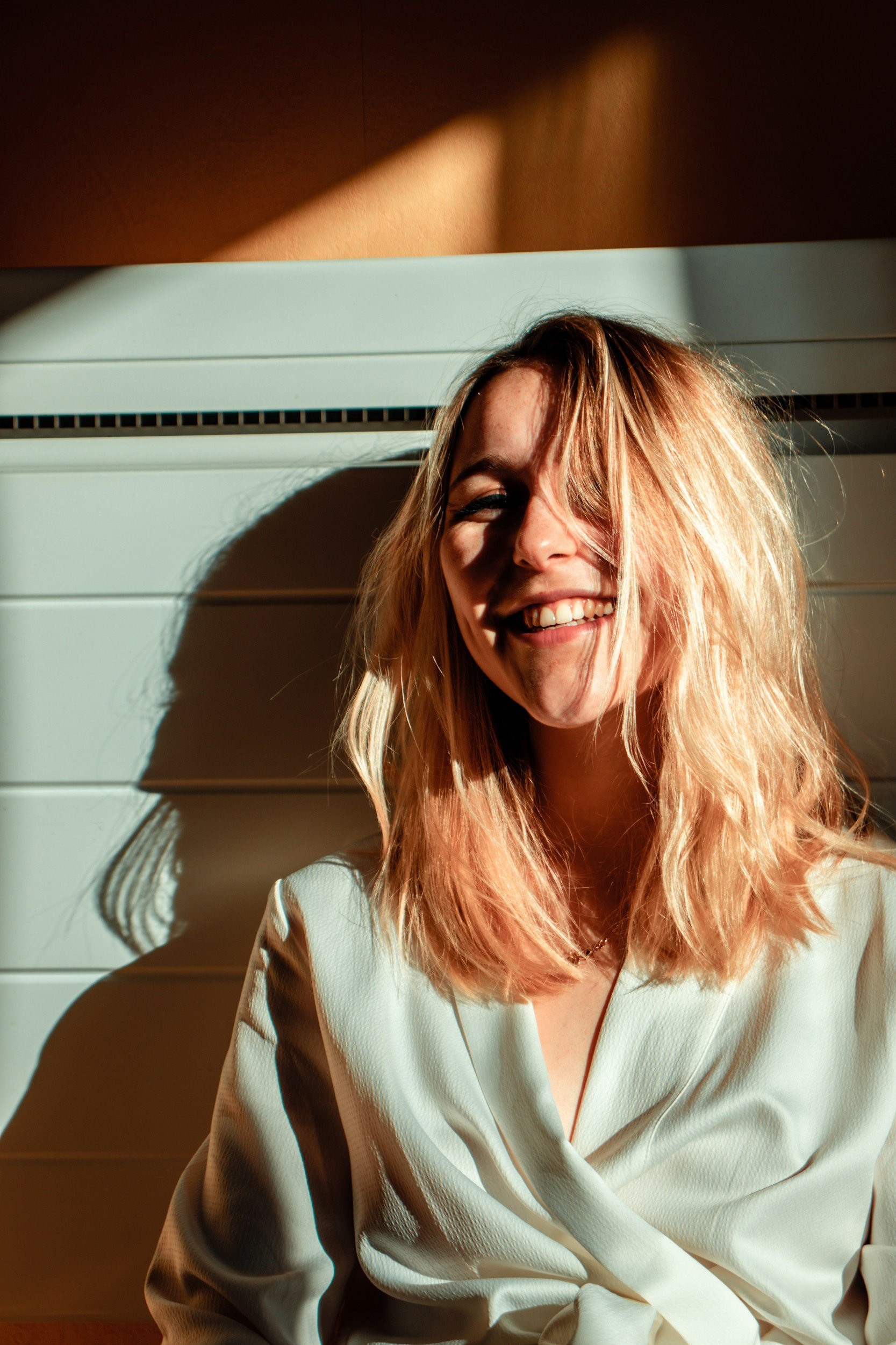 A woman with blonde hair smiling in sunlight, sitting against a white slatted wall.