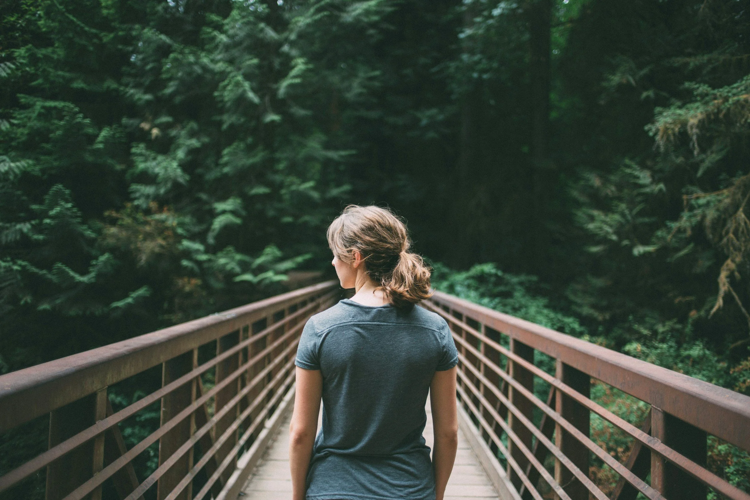 A woman with brown hair tied back in a ponytail, wearing a blue t-shirt, walking on a bridge through a lush green forest.