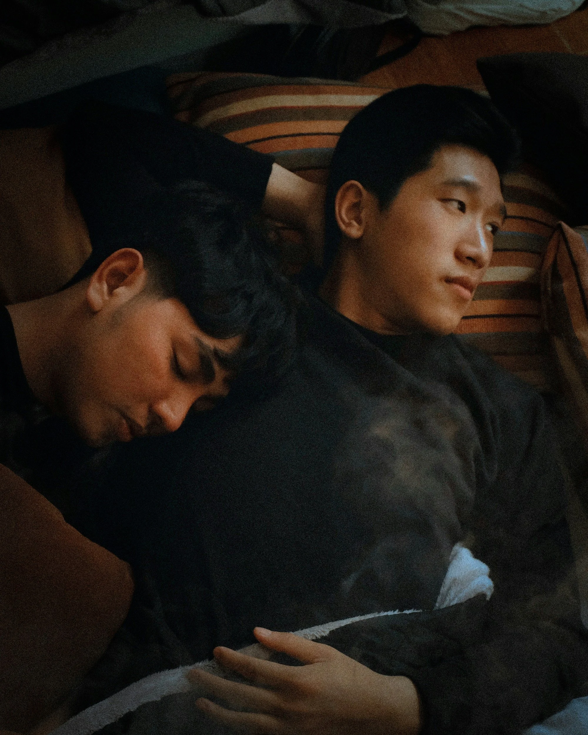 Two young people sleeping on a couch, with one resting their head on the other's arm. The person on the left has short dark hair and smooth skin, while the person on the right has short black hair and a soft expression. They are surrounded by striped pillows.