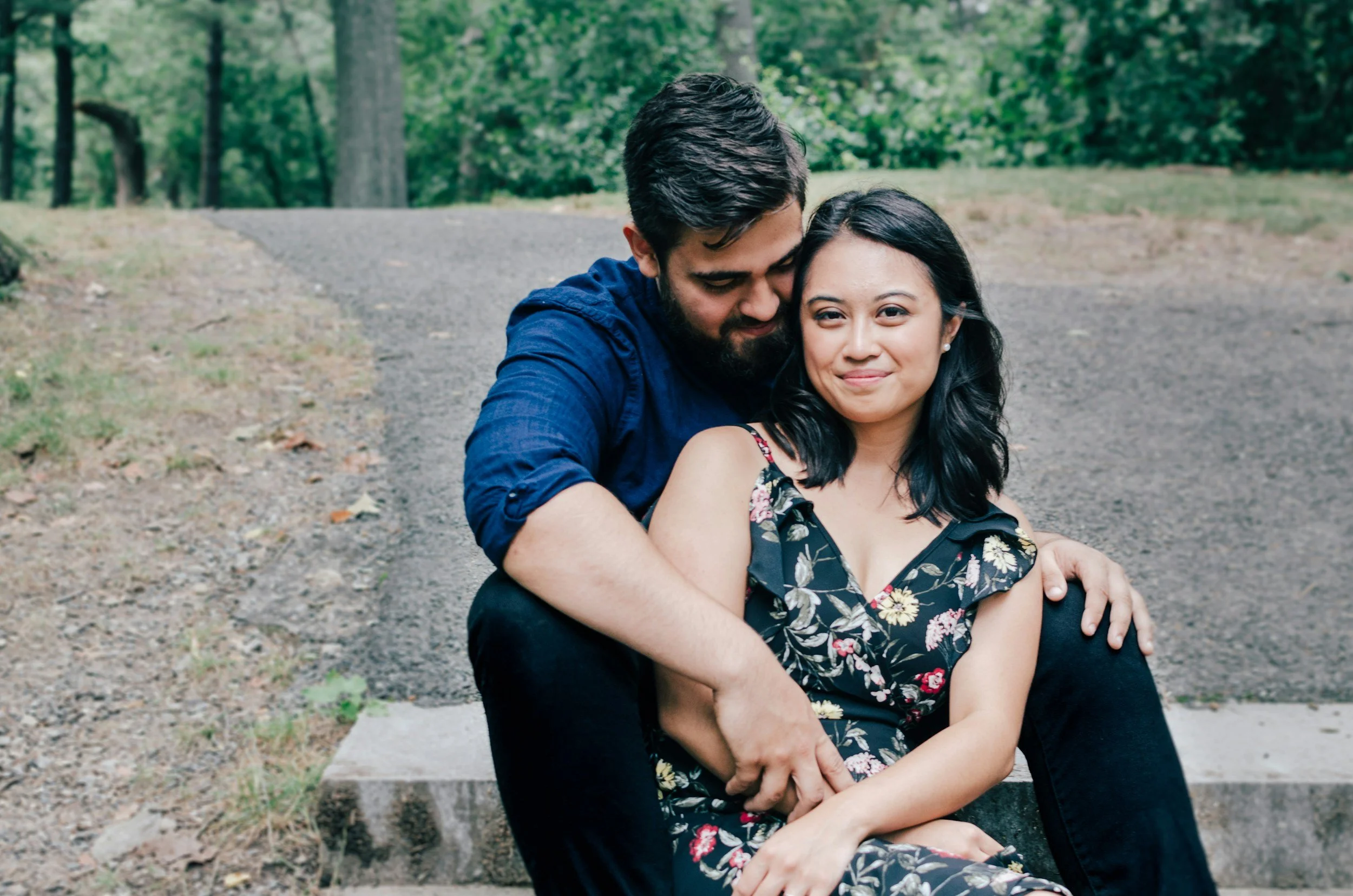 A couple sitting on a park bench, embracing each other with smiles. The man has dark hair and a beard, wearing a blue shirt. The woman has black hair and wears a floral dress. They are surrounded by trees and greenery.