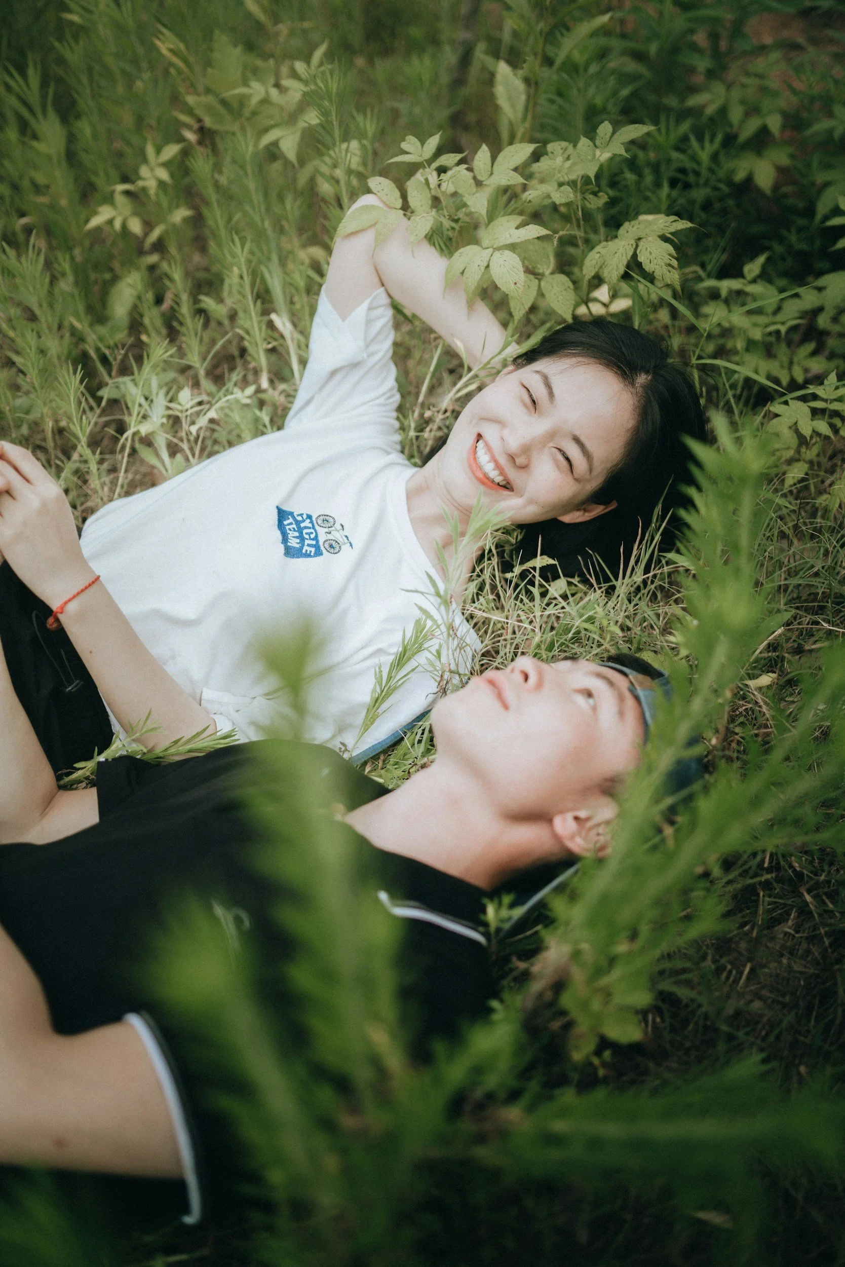 Two young people, a woman and a man, lying on the grass surrounded by green foliage, smiling and relaxed.