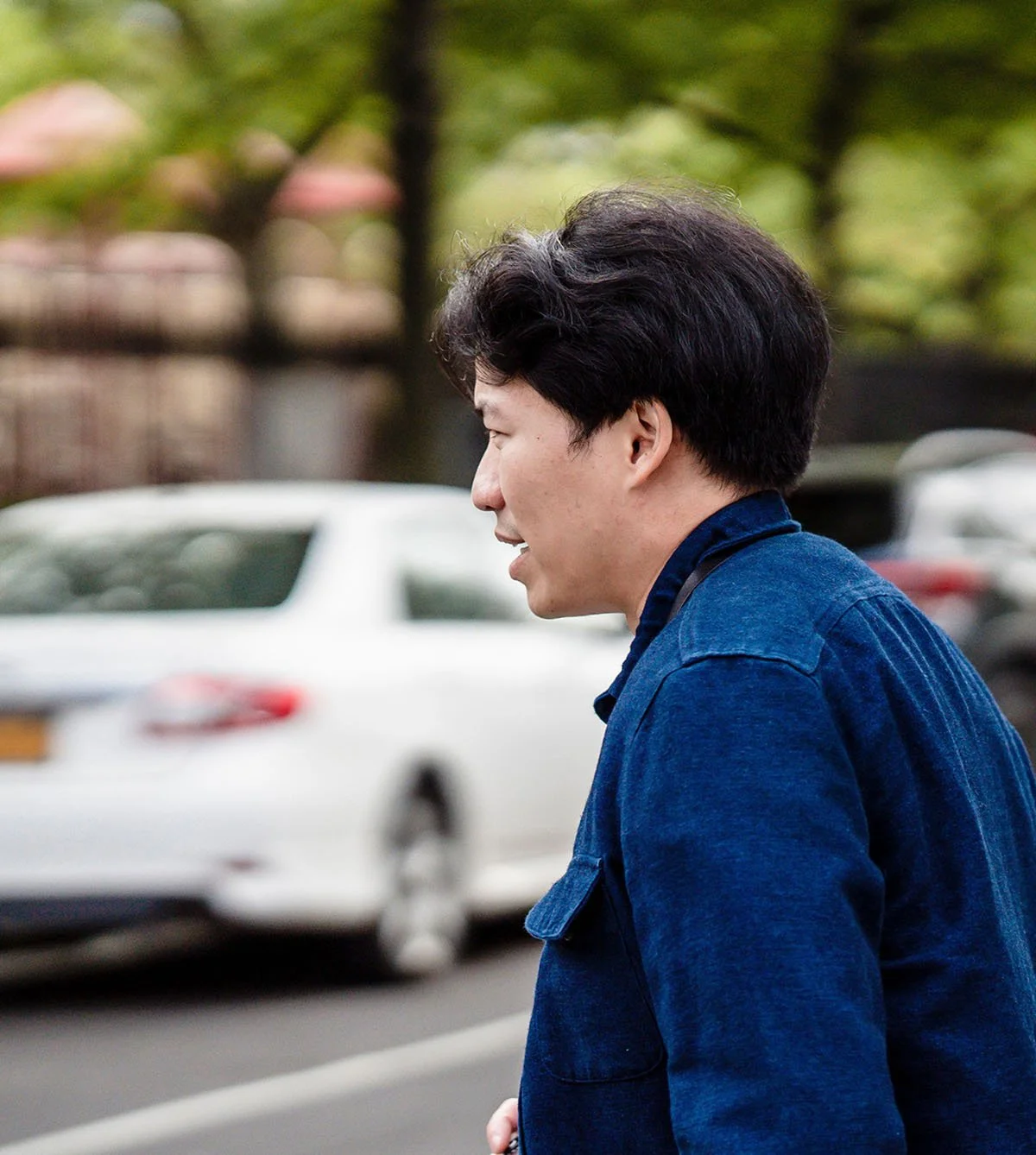 Side profile of a young man with dark hair wearing a denim jacket, walking outdoors with cars and trees in the background.