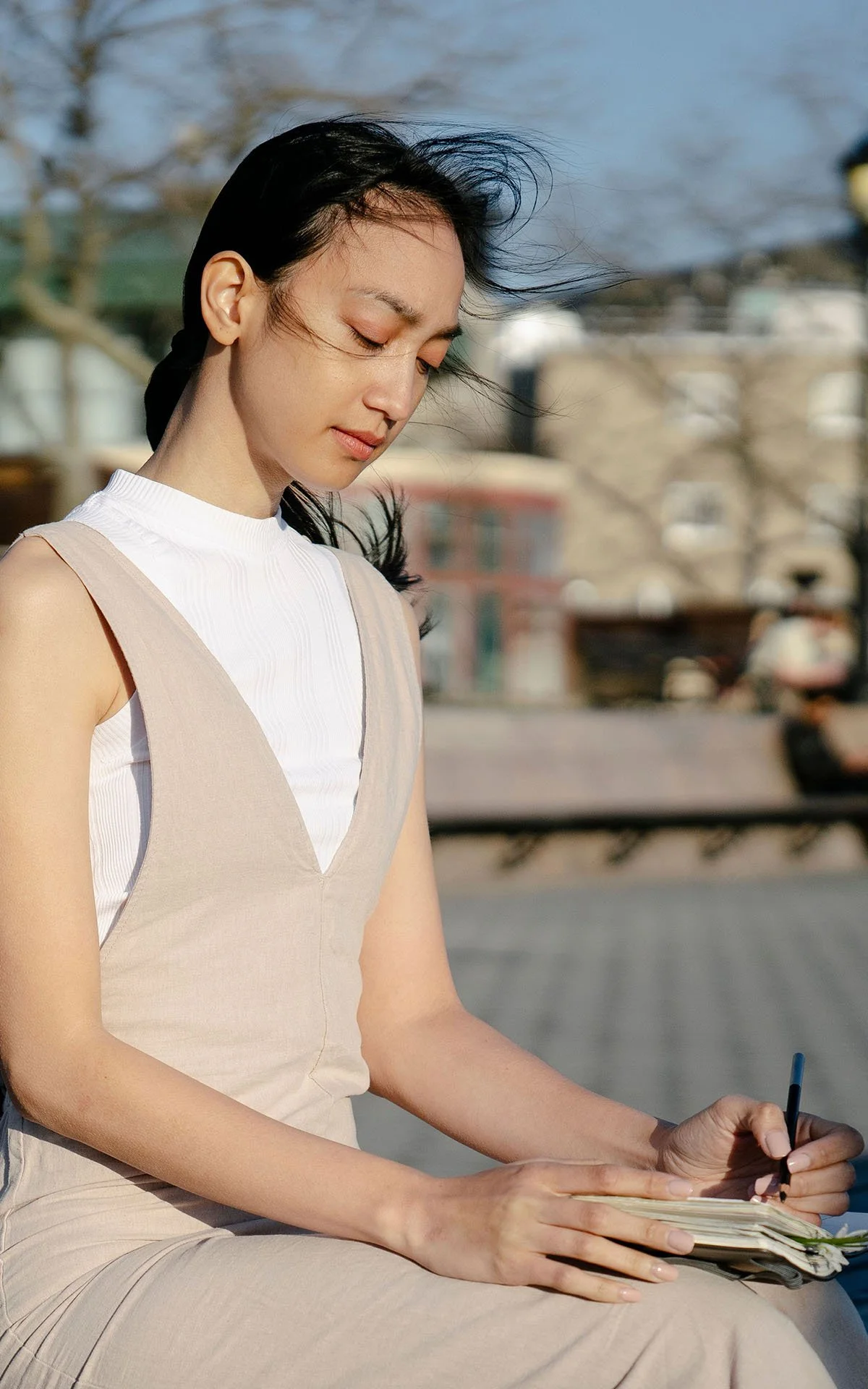 Young woman sitting outdoors, writing in a notebook, with wind blowing her hair.