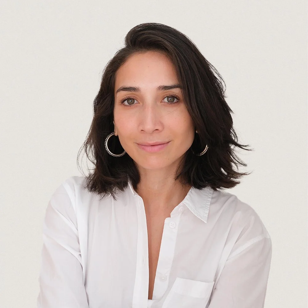 A woman with shoulder-length wavy dark hair wearing a white button-up shirt and hoop earrings.