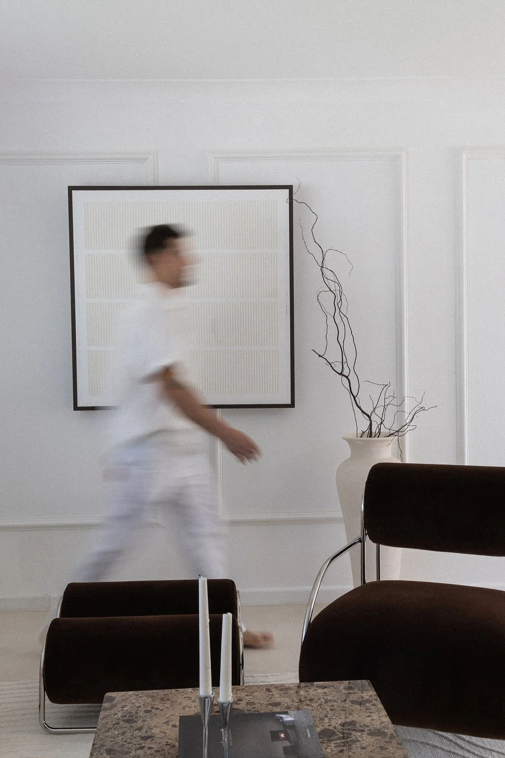A blurred person walking past a modern living room with white walls, a framed piece of art, a tall white vase with twisted branches, and minimalistic furniture including a black and brown chair and a marble table with candles.