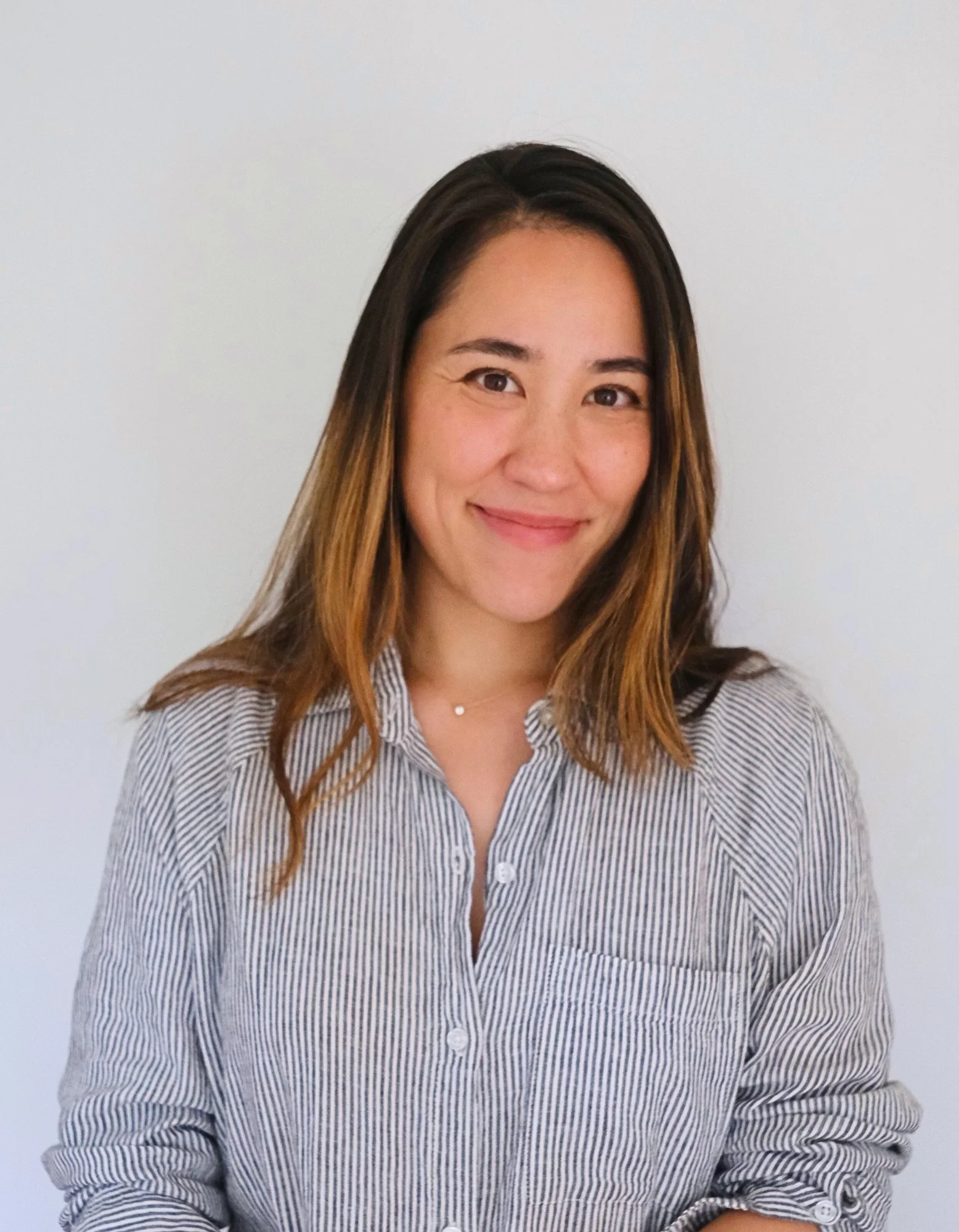 A woman with shoulder-length brown hair, wearing a striped button-up shirt, smiling against a plain white background.