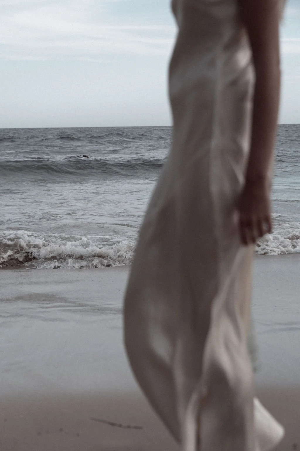 A person in a beige dress standing on a sandy beach near the ocean with waves, with the ocean and cloudy sky in the background.
