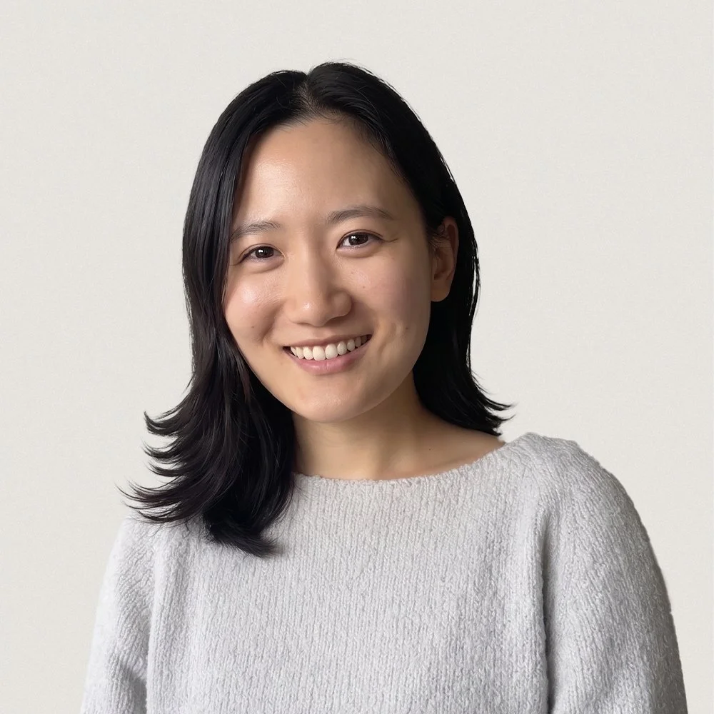 Portrait of a woman with black hair smiling, wearing a light gray sweater, standing against a plain light-colored background.