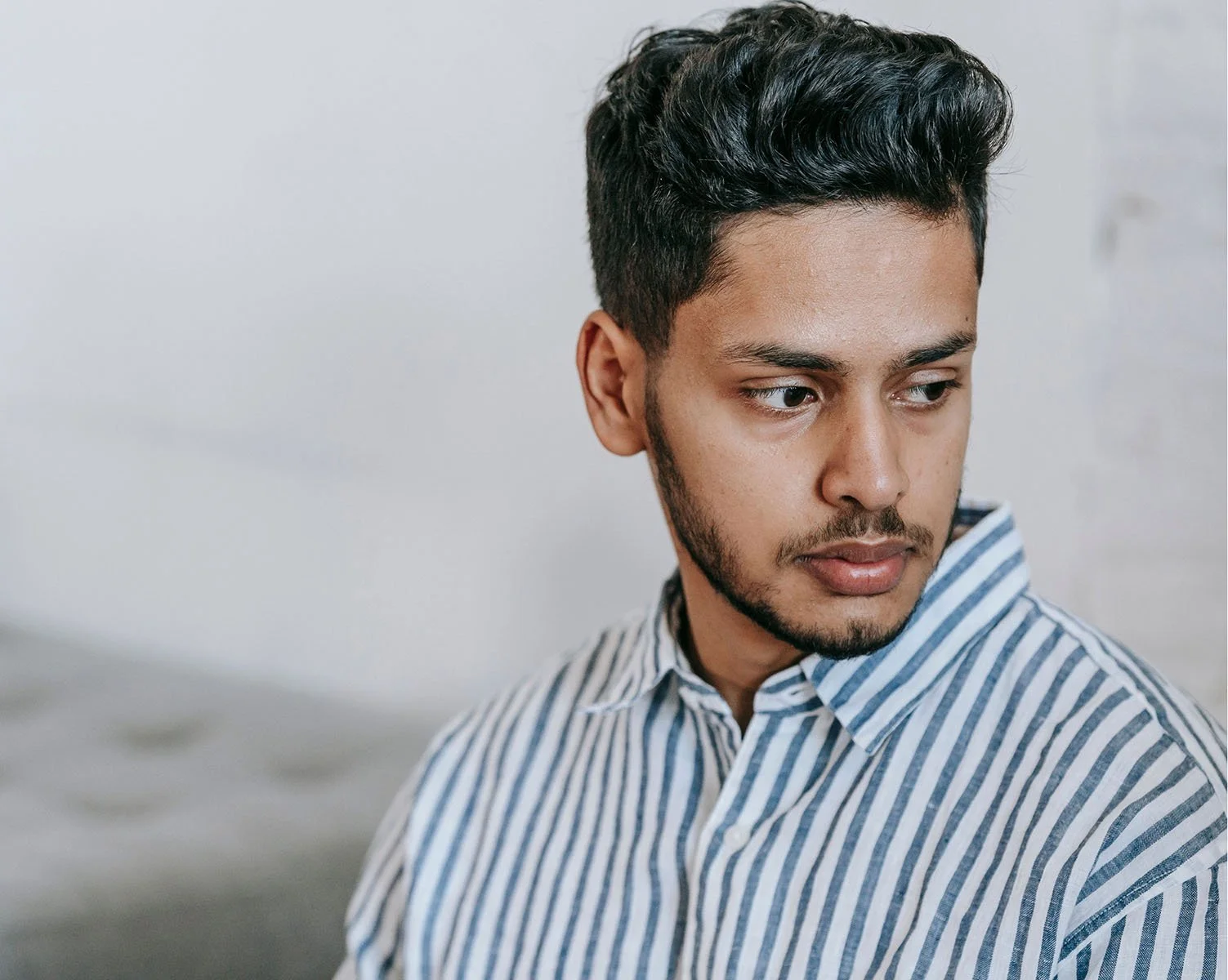 A young man with dark hair and a beard looking to the side, wearing a striped shirt, against a plain white background.