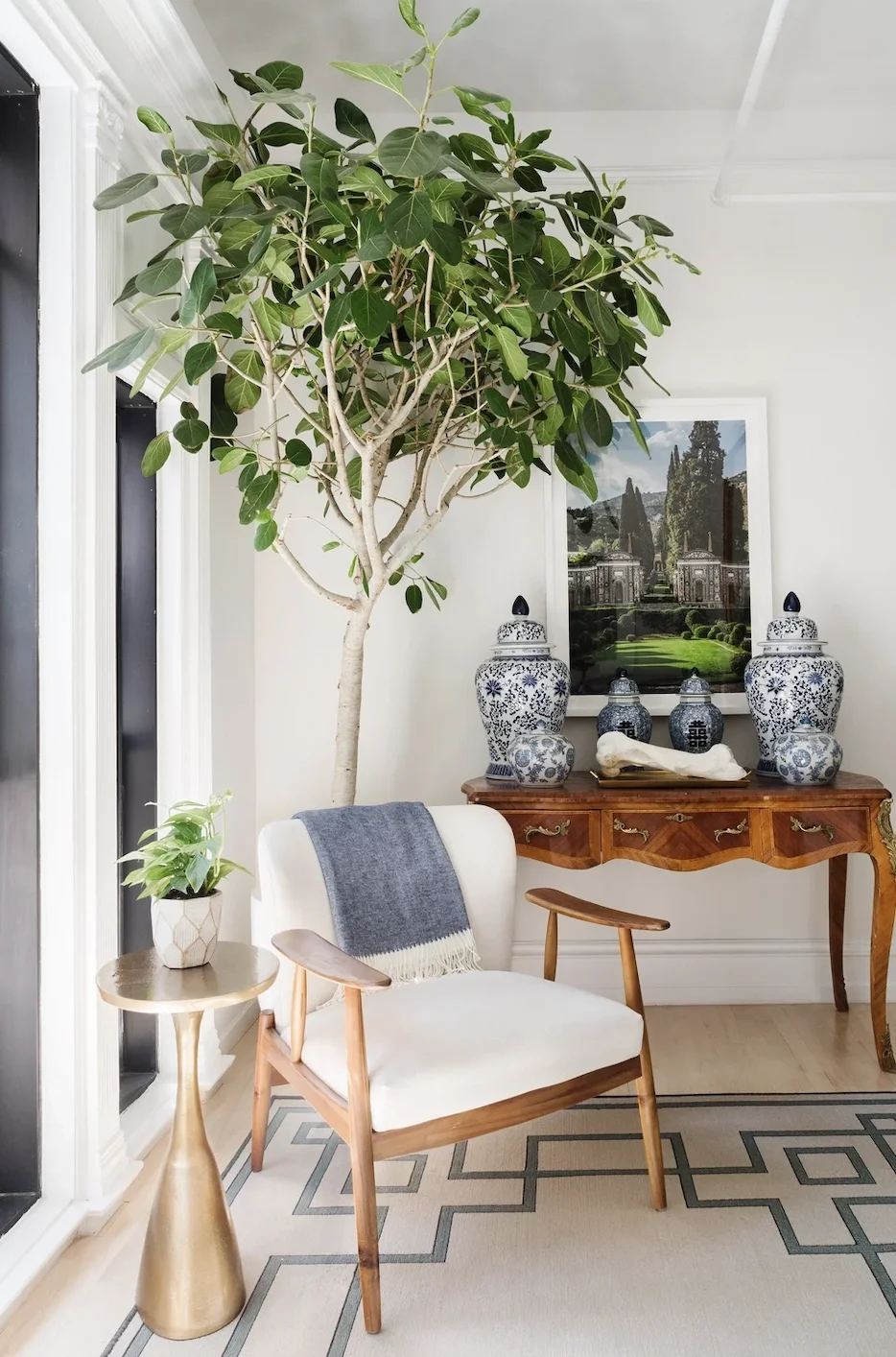 A cozy reading nook with a large potted tree, a white armchair with wooden armrests and a gray throw blanket, a small side table with a potted plant, a wooden console with blue and white vases, and framed gallery wall art in a bright room.