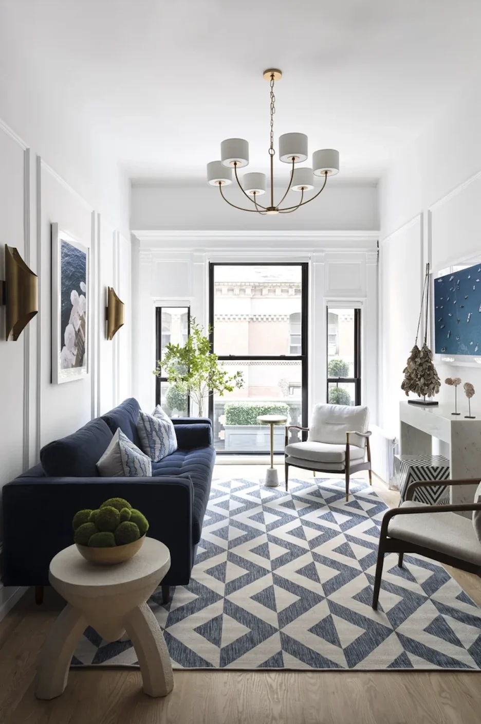 A modern living room with a white ceiling, a chandelier, black-framed windows, a navy sofa with pillows, a white armchair, a geometric patterned rug, and decorative plants and wall art.
