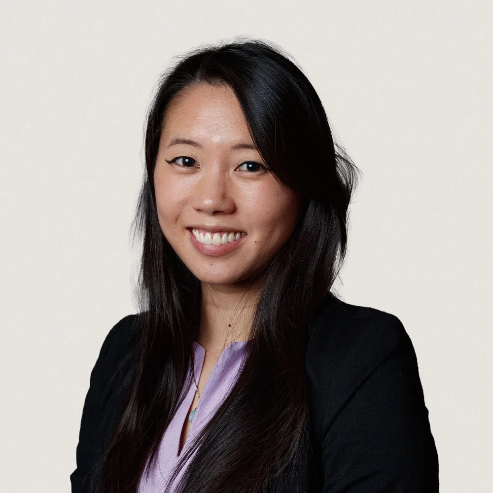 A young woman with straight black hair, smiling, wearing a black top and a purple shirt underneath, against a plain white background.