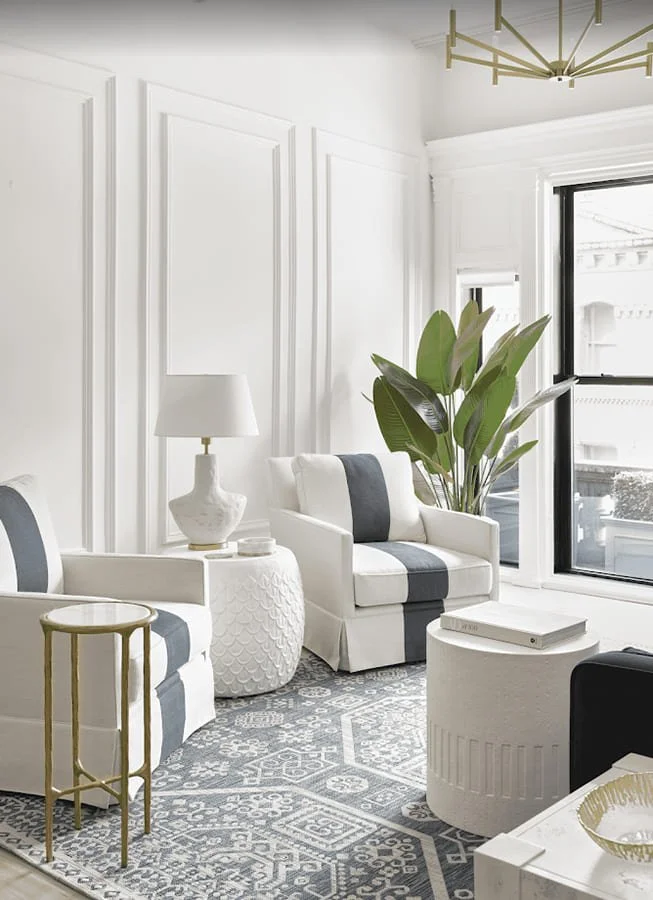 Bright living room with white walls, large window, white and gray furniture, a potted plant, a side table with a lamp, and a patterned rug.