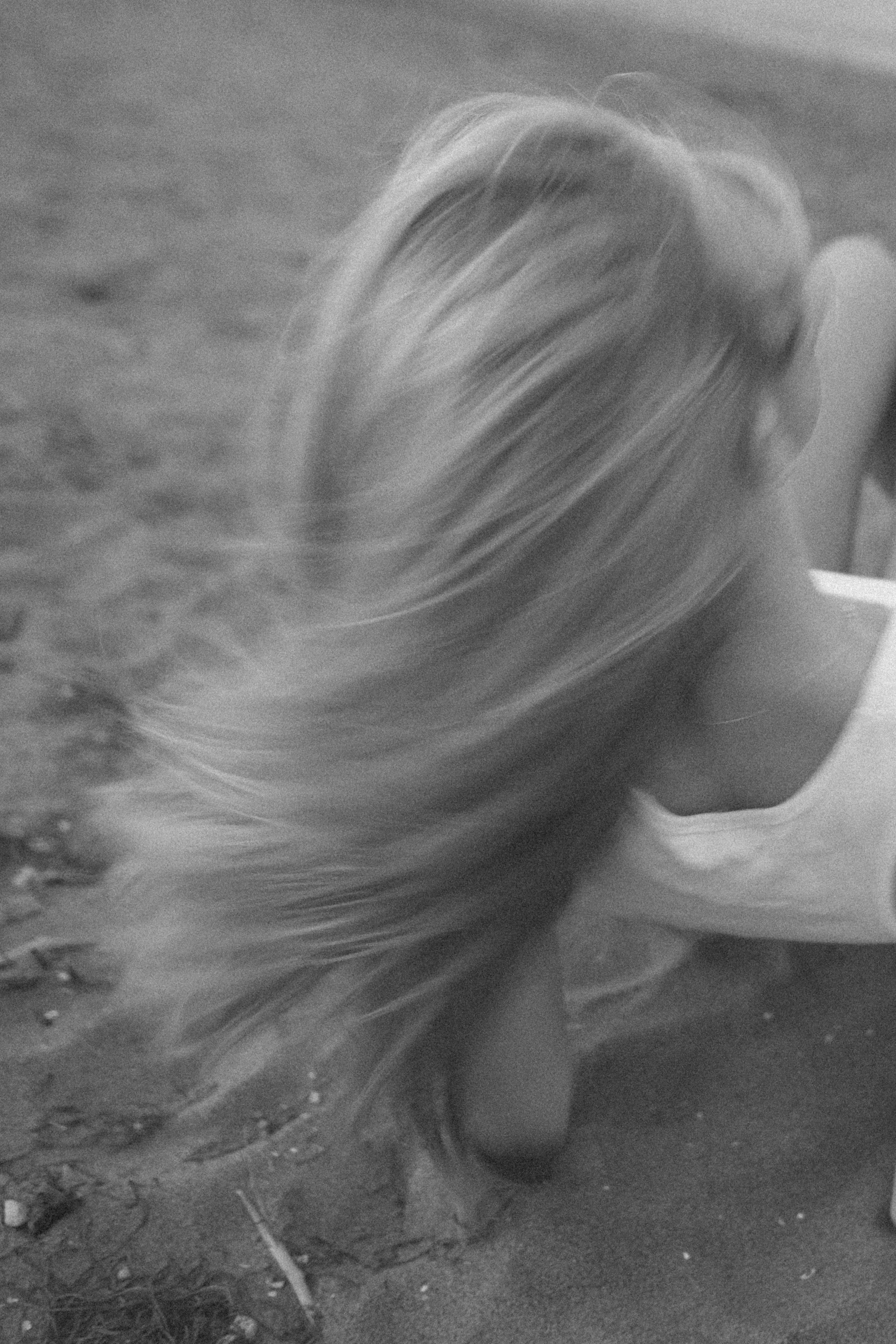 A woman with long, flowing hair, shown from the side, is kneeling on a sandy surface. She is wearing a mask and casual clothing.
