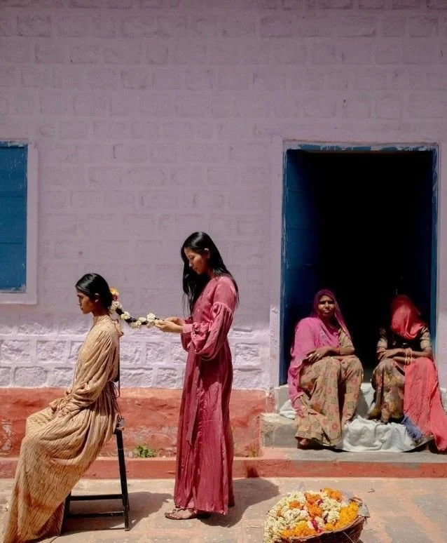 A girl sits on a chair while another girl stands behind her, holding a floral garland. Two women sit outside a blue doorway in the background.