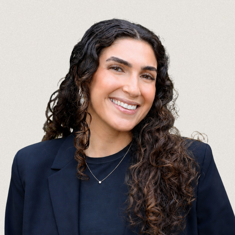 A woman with long curly brown hair, smiling, wearing a black blazer and a delicate necklace, standing against a plain, light-colored background.