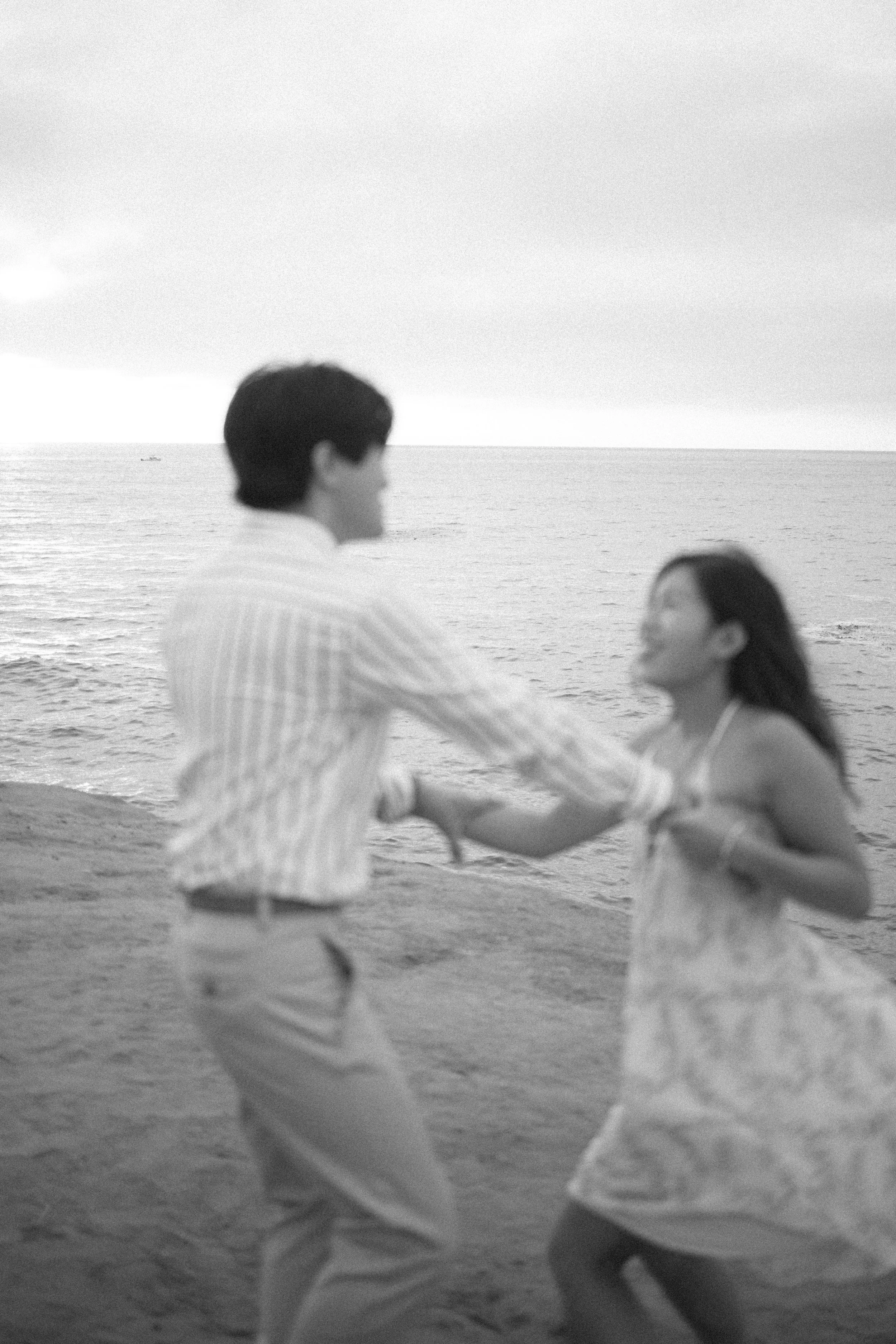 Black and white photo of a man and woman holding hands on the beach, with the ocean in the background.