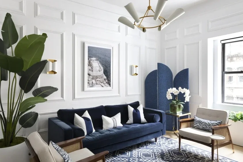 Living room with white paneled walls, a navy blue sofa with white and navy pillows, a white armchair with a zebra print pillow, a dark blue room divider, a large window, a patterned rug, and a chandelier with white shades.