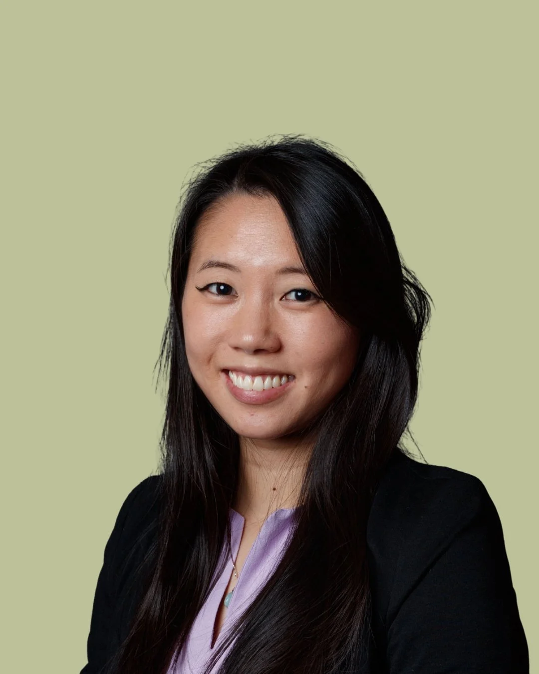 A professional woman with long dark hair, smiling, wearing a black blazer and a light purple blouse, against a light green background.