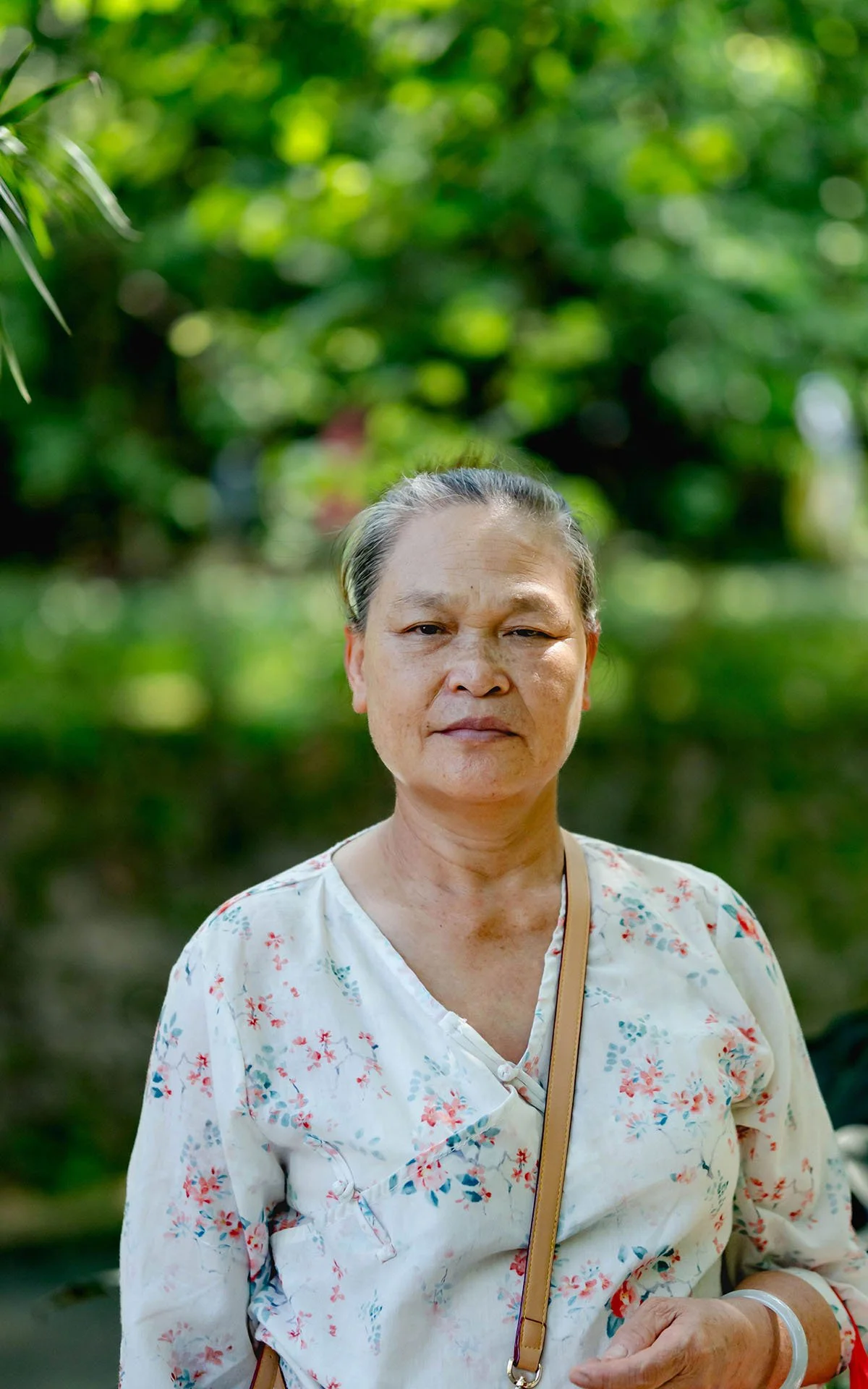 An older woman with short gray hair, wearing a white floral blouse, stands outdoors with a blurred green leafy background.