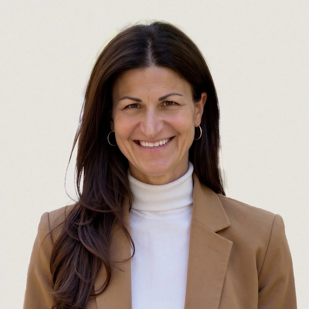 A woman with medium-length dark hair, wearing hoop earrings, a white turtleneck, and a light brown blazer, smiling in front of a plain white wall.