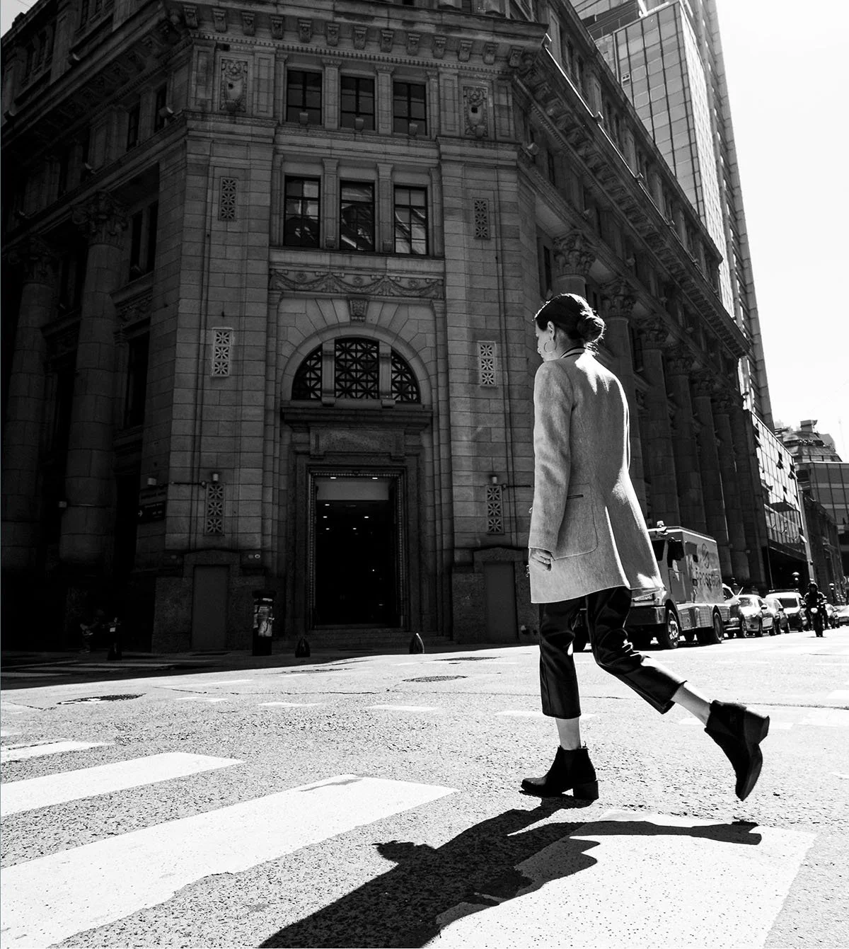 A woman crossing a city street at a crosswalk in front of a large historic building, captured in black and white.