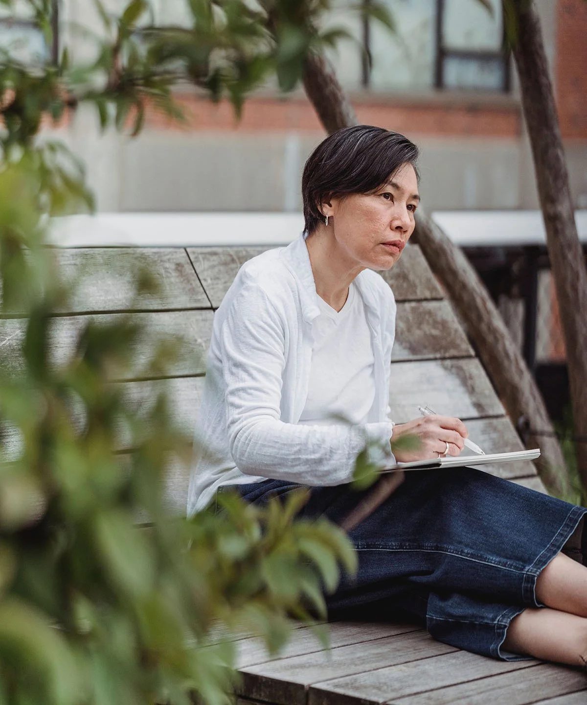 A woman sitting on a wooden bench outdoors, writing in a notebook with a pen, surrounded by trees and greenery.
