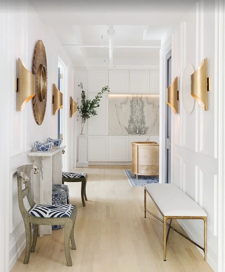 A bright, modern hallway with white paneled walls and light wood flooring. Decor includes gold wall sconces, a large woven wall art piece, a small white console table with blue and white bowls, two chairs with striped seats, a gold and white bench, and a large green plant in a pot. In the background, a built-in white cabinet and a marble accent wall with a vase and branches are visible.